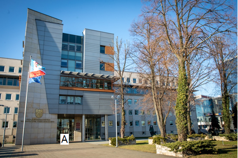 The main four-story building of the Marshal's Office in Rzeszów at ul. Cieplińskiego 4. Bright facade. The facade of the building in a modern style rises upwards. The glass door illuminates the entrance. On the front wall there is a golden coat of arms of the Podkarpackie Region. The griffin crowned on it symbolizes the Bełz Region. Next to it, a crowned lion symbolizes the Lviv Region. Above them is a knight's cross. Under the coat of arms, a golden inscription: Marshal's Office of the Podkarpackie Region. In front of the building there is a flag with the coat of arms of the Podkarpackie Region, the flag of Poland and the flag of the European Union. Trees and bushes on the right side of the building. Behind them is a monument to the victims of the Smolensk crash symbolizing the plane's wing damaged during the flight. Next to it, there is another monument - a stone commemorating the 1050th anniversary of the Baptism of Poland.