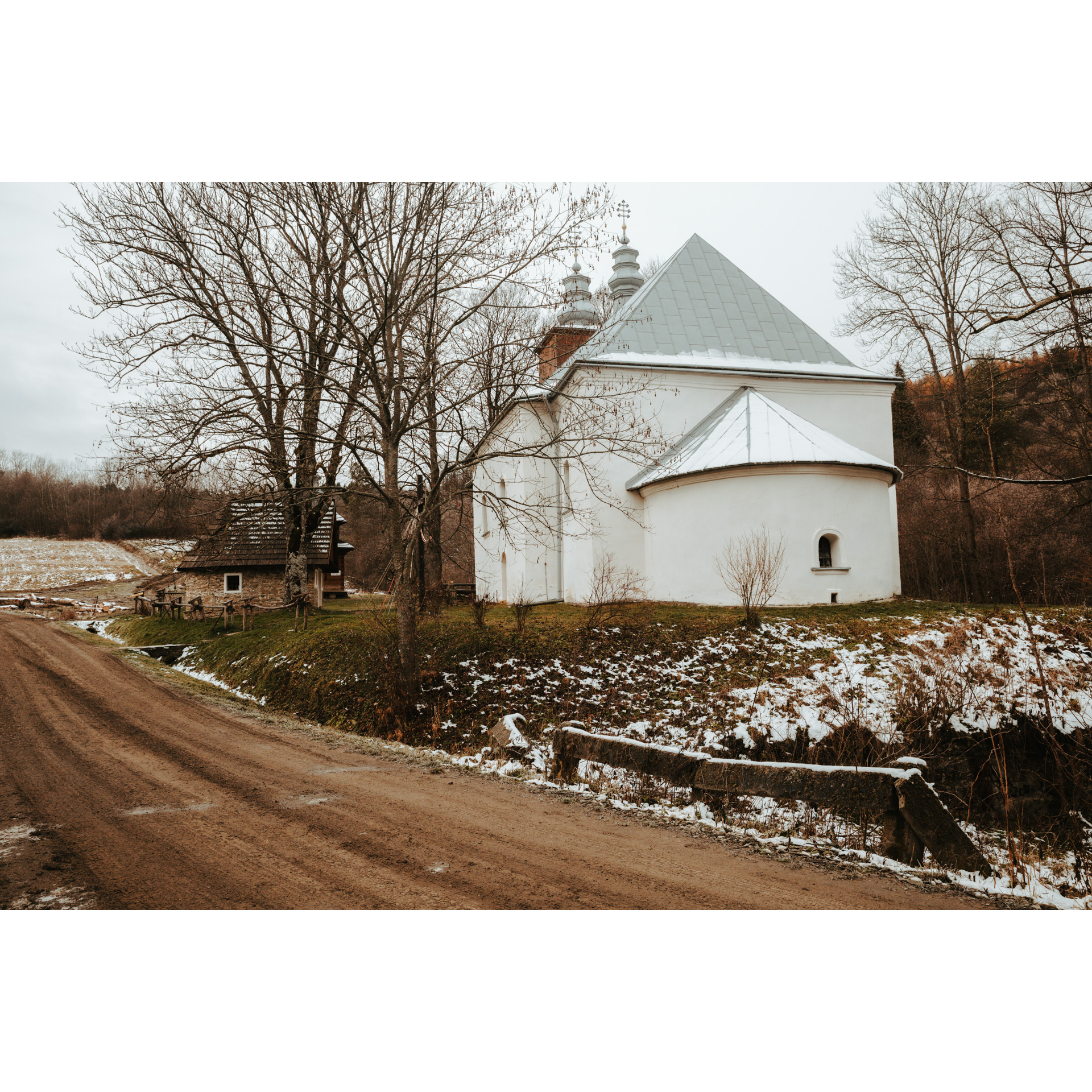 A white church building by a dirt road and an old house with a wooden roof