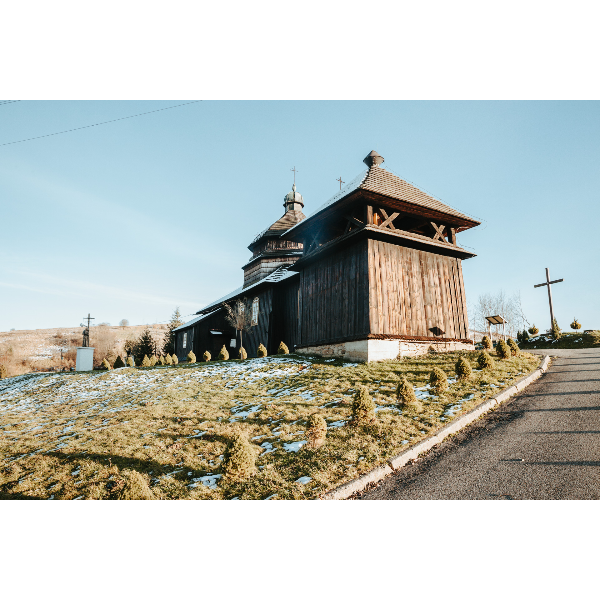Drewniana cerkiew na trawiastym wzgórzu obok krzyża i szerokiej drogi prowadzącej do obiektu