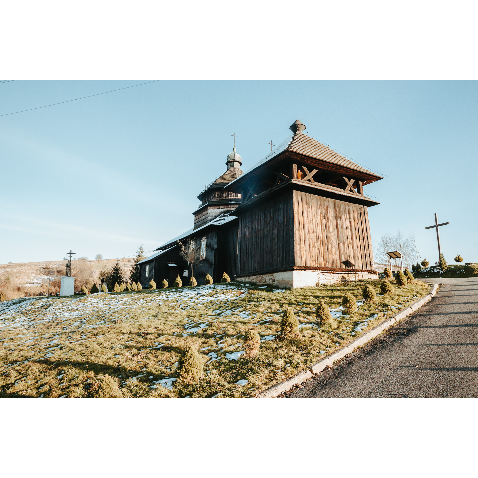 A wooden church on a grassy hill next to a cross and a wide road leading to the facility