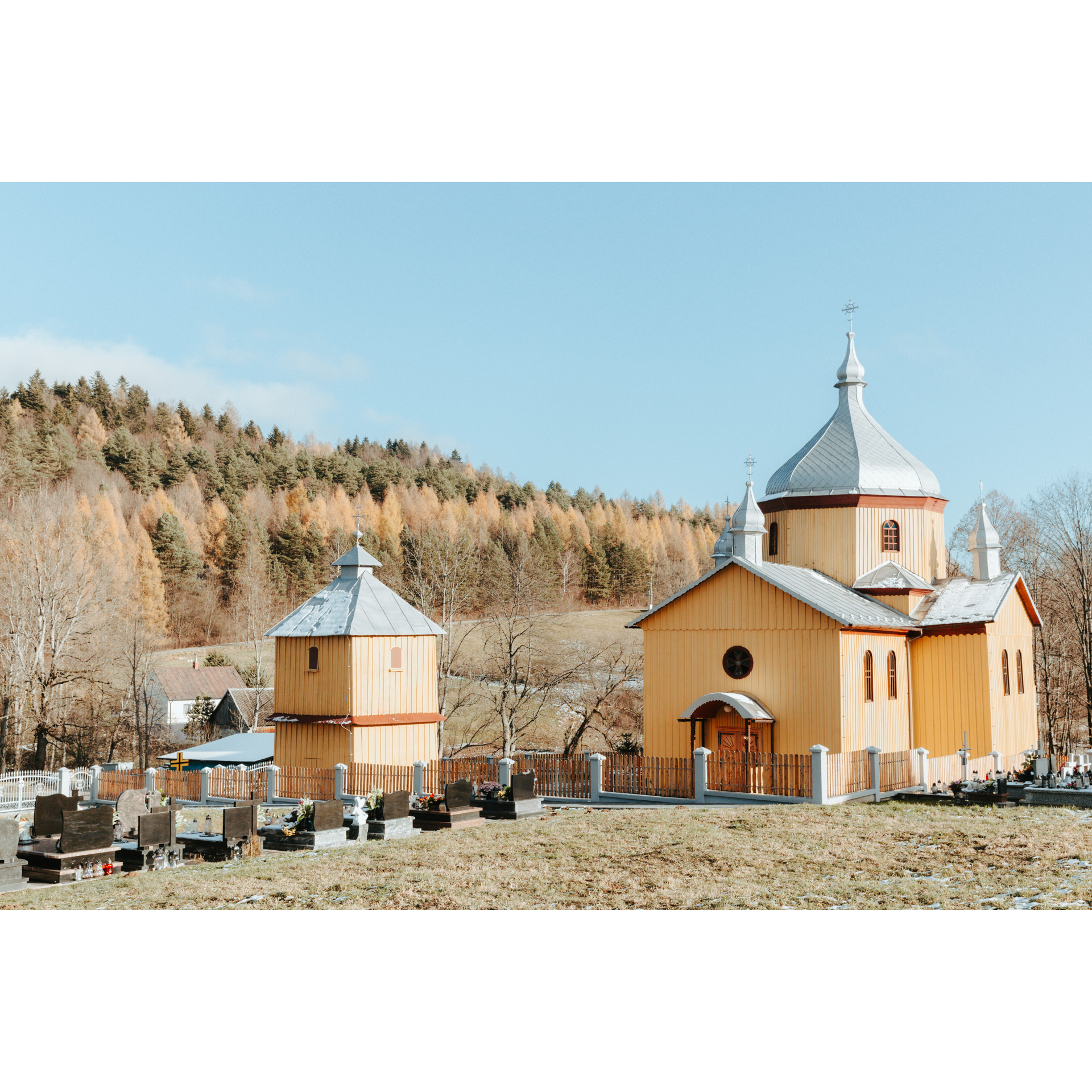 Drewniana cerkiew o szarym dachu z dzwonnicą i cmentarzem na przodzie