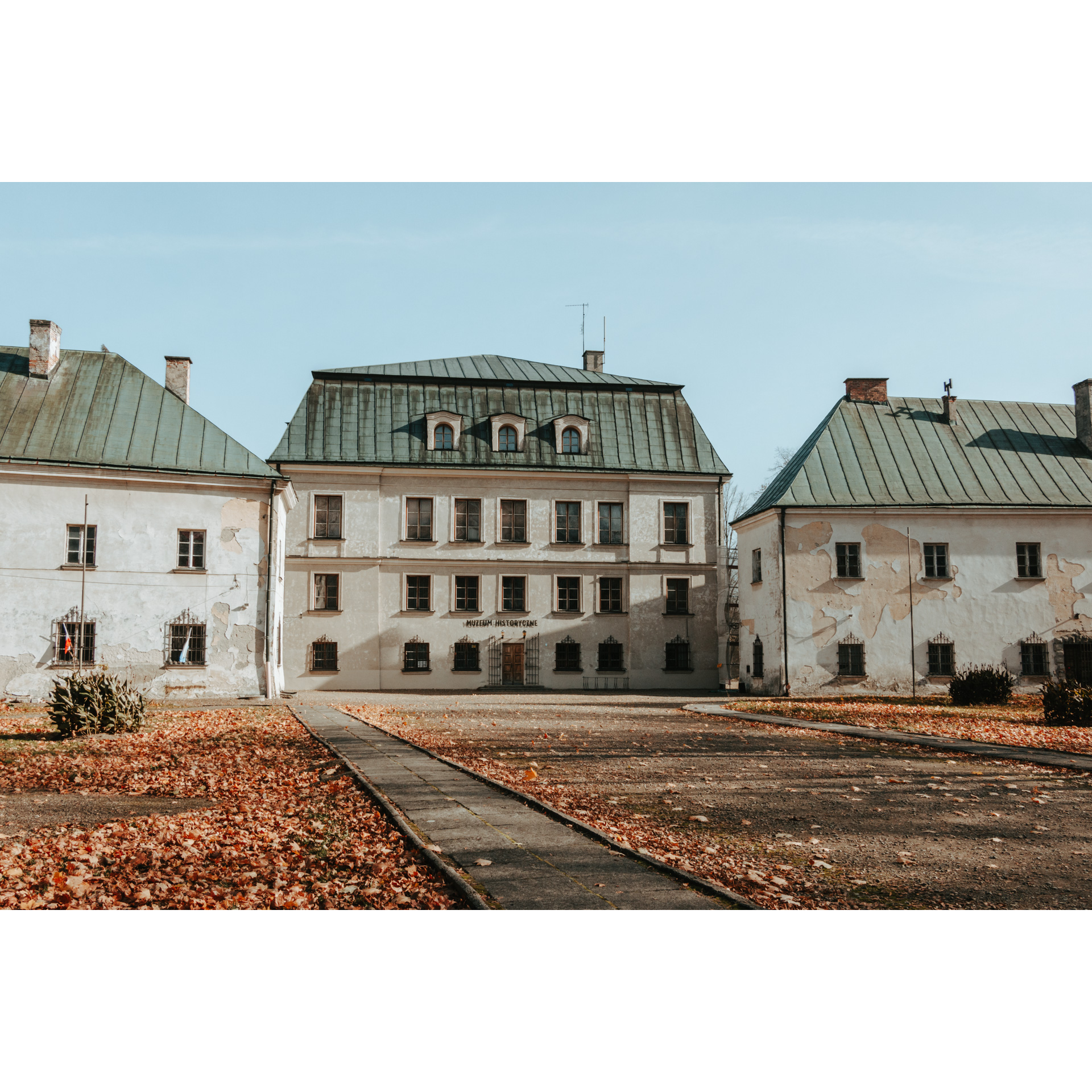 Trzy nieco zniszczone murowane, kilkupiętrowe jasne budynki z zielonym dachem stojące obok siebie