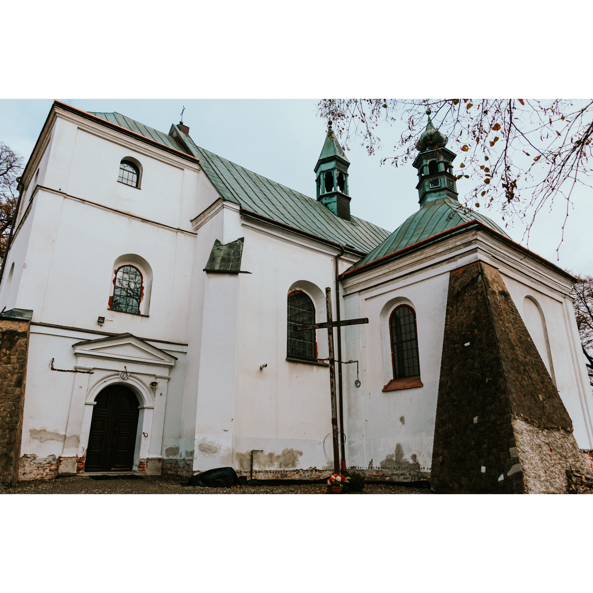 Jasny, murowany kościół z zielonym, spadzistym dachem i czarnymi półokrągłymi drzwiami obok wysokiego, czarnego krzyża
