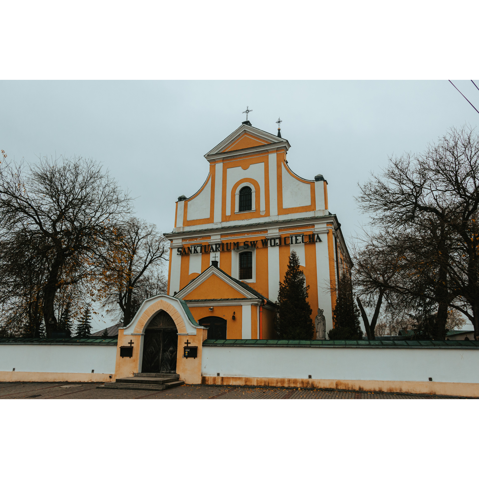 Biało pomarańczowy kościół z ozdobną fasadą i czarnym napisem ogrodzony jasnym murem z pomarańczową murowaną bramą