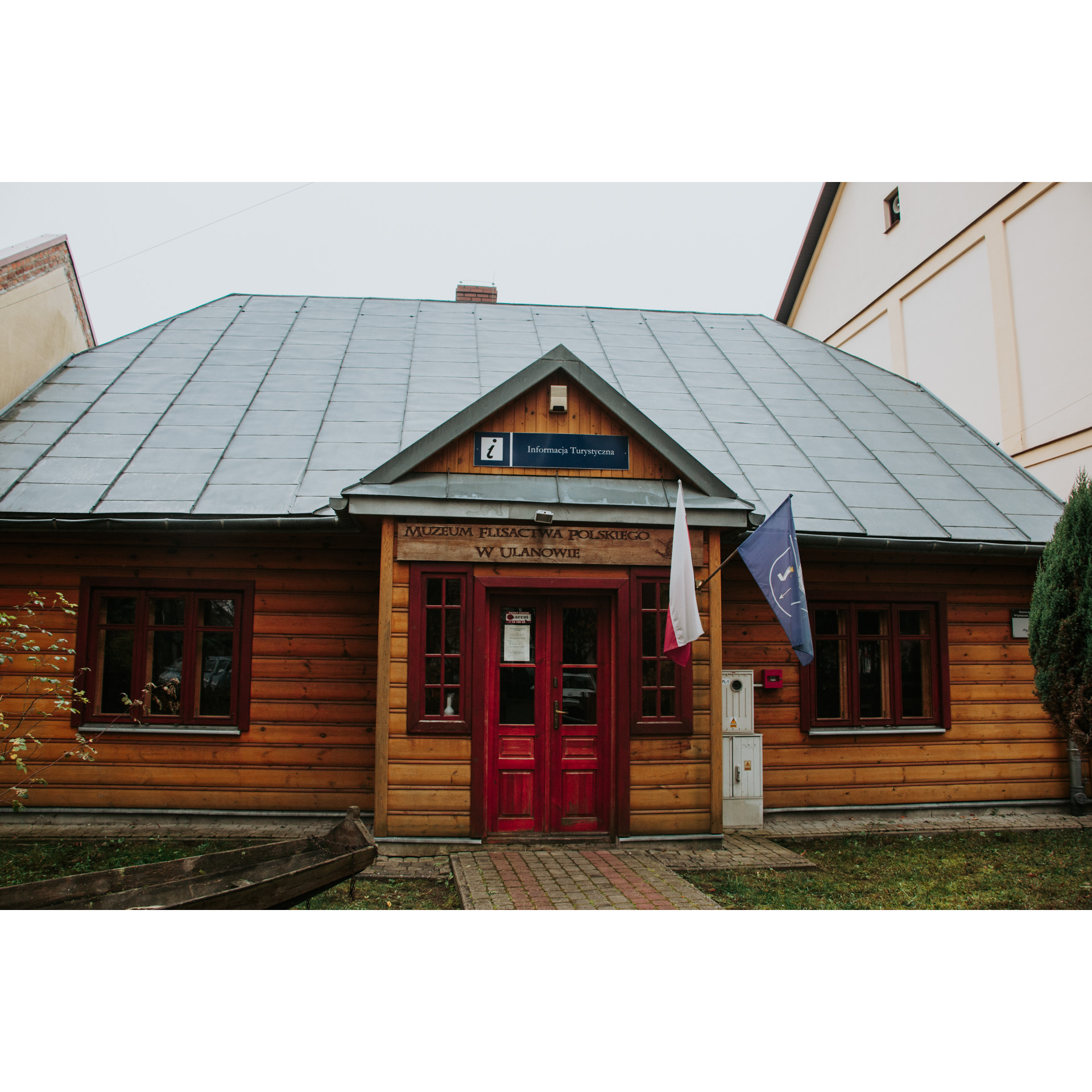 Wejście do drewnianego budynku muzeum z czerwonymi, oszklonymi drzwiami i szarym dachem