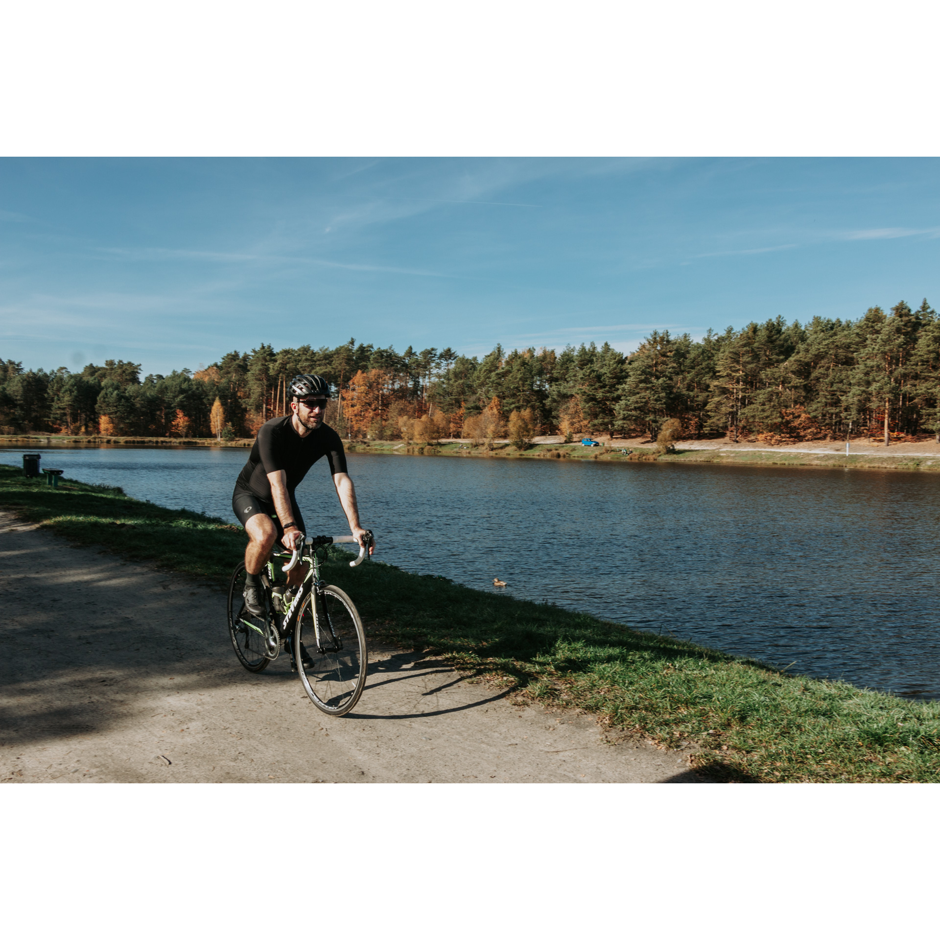 A cyclist in a black outfit and a helmet riding on a road along a river with a forested shore