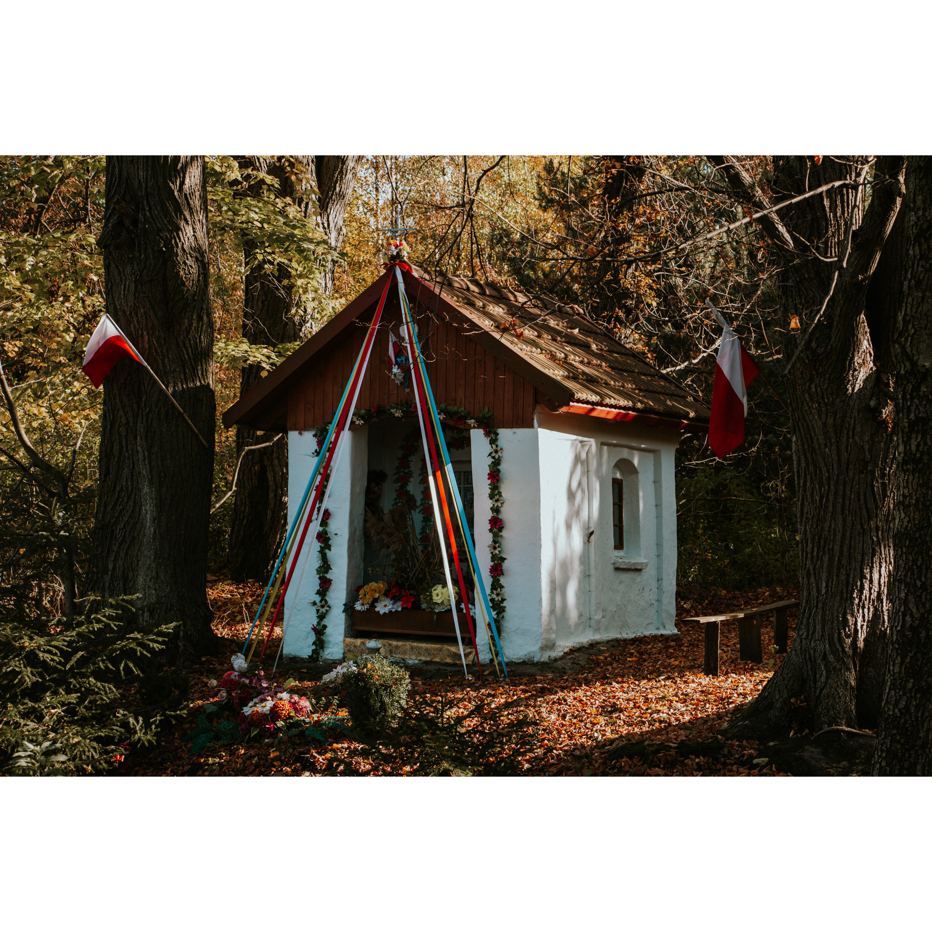 A white chapel in the forest, with a wooden roof, decorated with flowers and colorful ribbons and Polish flags