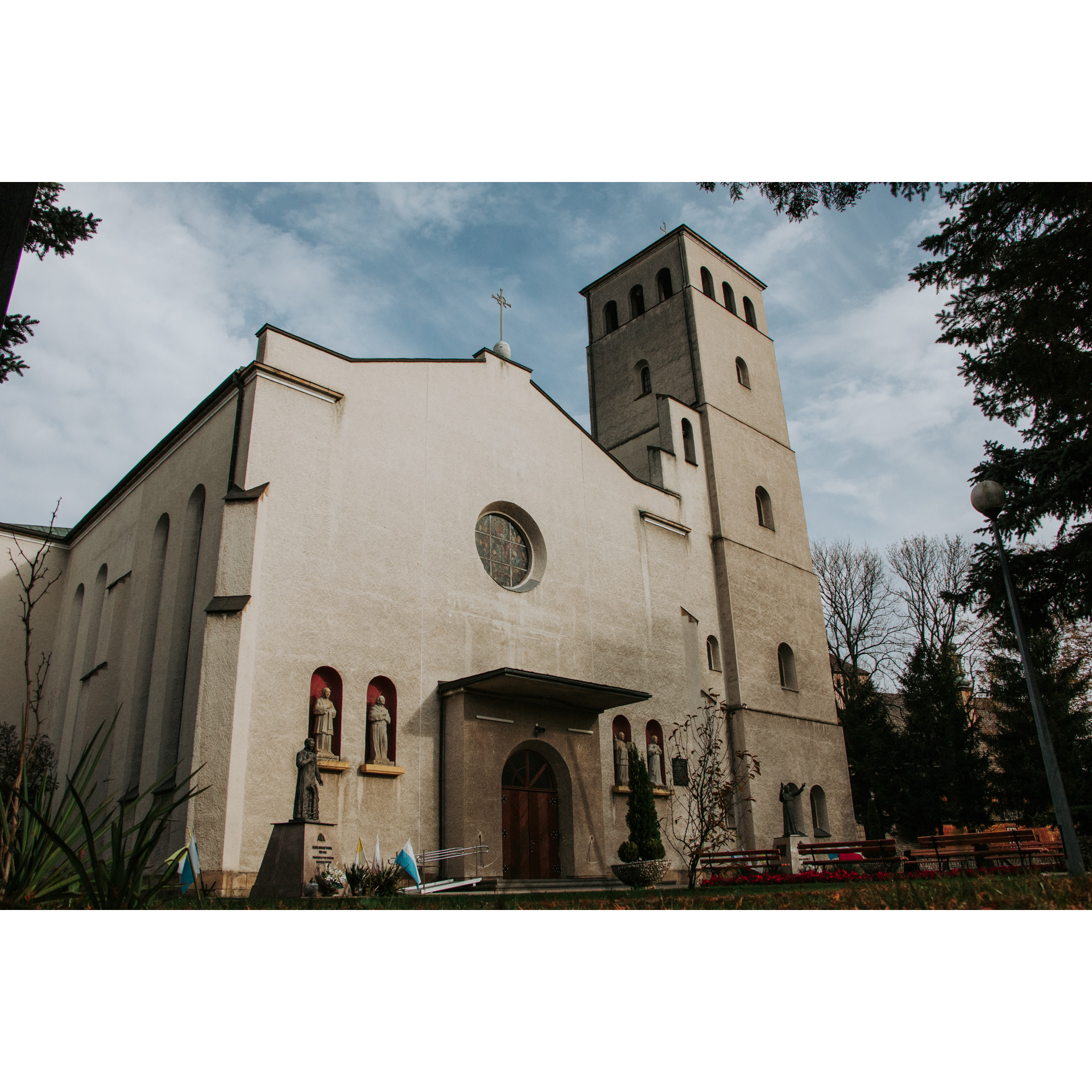 A brick church in gray with a very wide facade and a high tower, on both sides of the entrance there are figures of saints