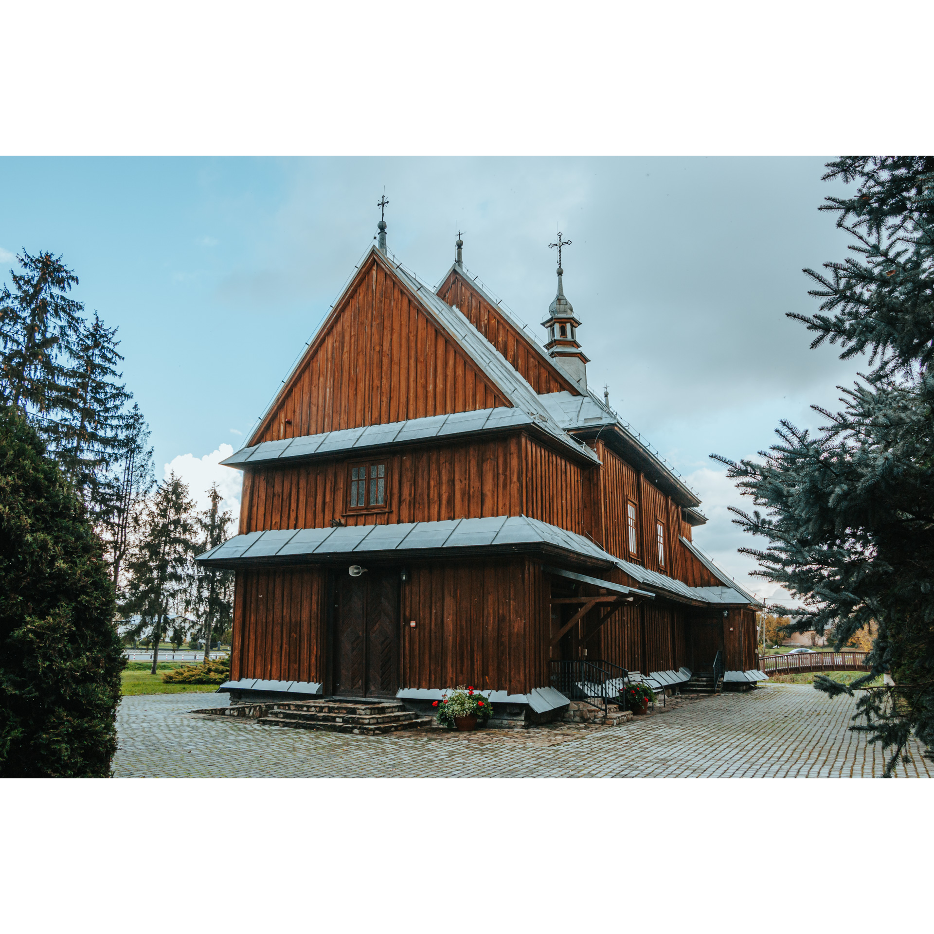 A wooden building made of vertical brown boards with a metal roof topped with small domes, a wooden dark door in the center, coniferous trees around