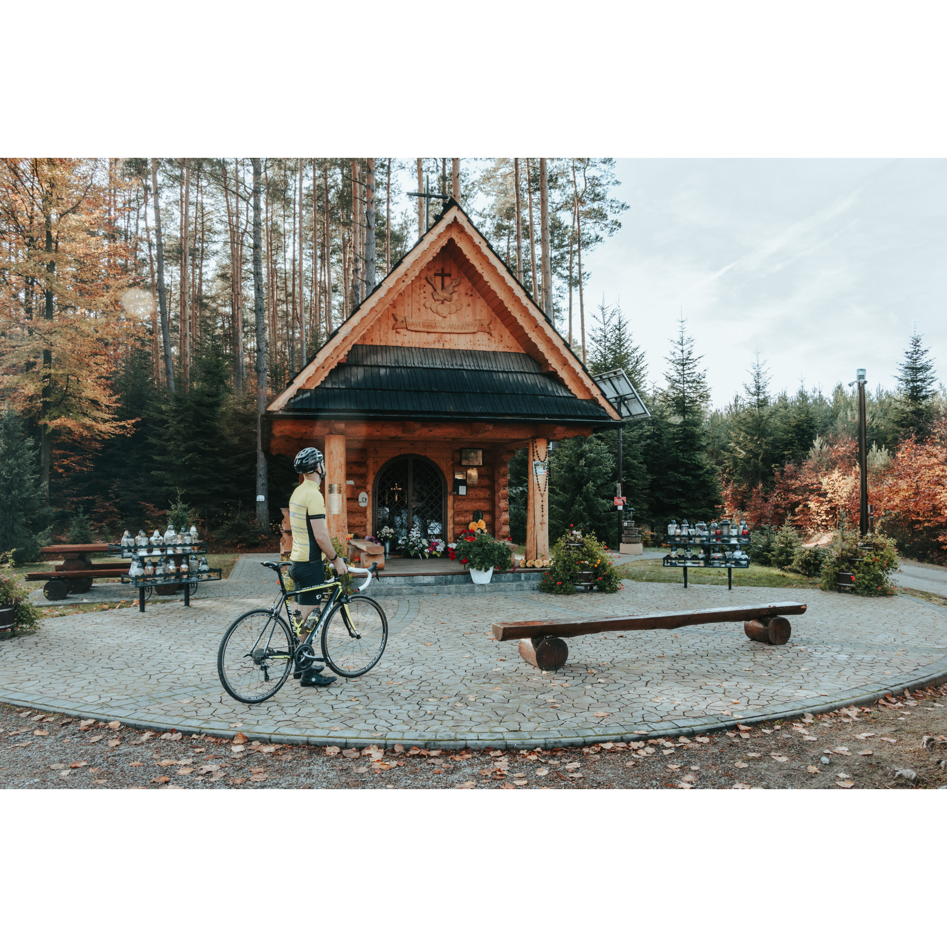 Drewniana kapliczka pośrodku jesiennego lasu umiejscowiona na wybrukowanym małym placu, wokół proste, drewniane ławki, a także znicze i kwiaty, w centrym rowerzysta w żółtym stroju sportowym, kasku, trzymający rower