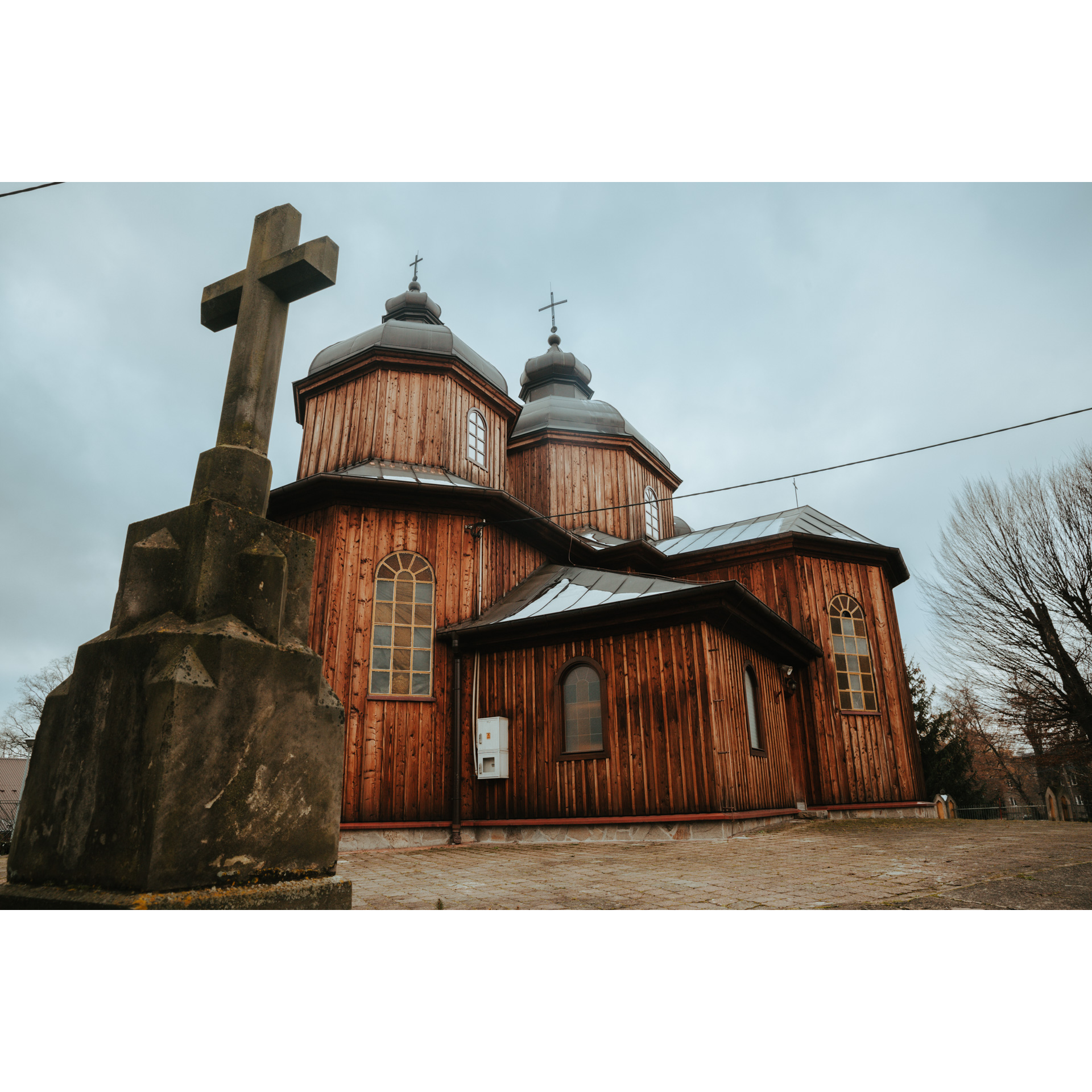 Drewniana cerkiew wykonana z desek pionowych, z dachem pokrytym metalem, z dwoma kopułami na tle nieba pokrytego deszczowymi chmurami. Na pierwszym planie kamienny krzyż