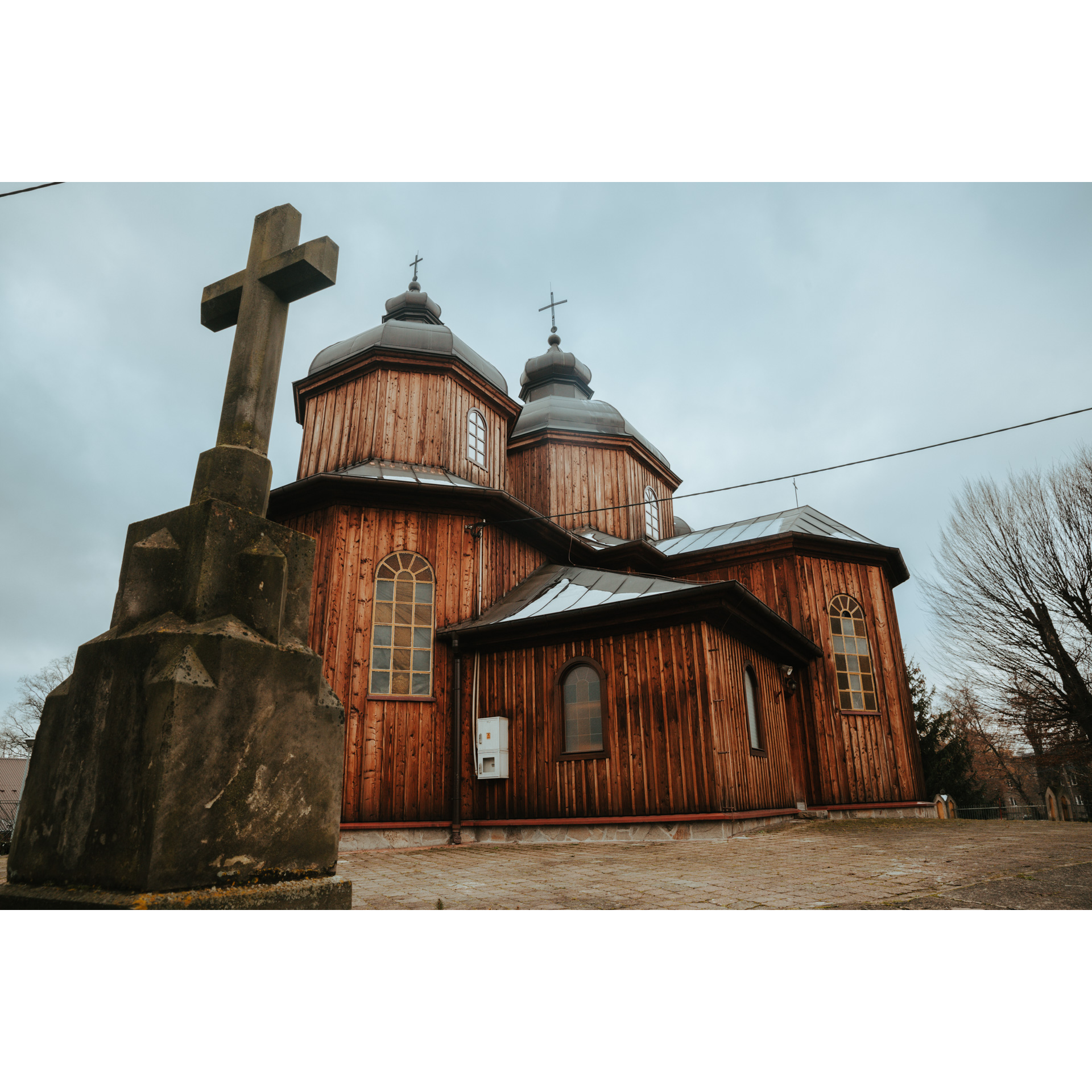 A wooden church made of vertical boards, with a roof covered with metal, with two domes against the sky covered with rain clouds. In the foreground a stone cross