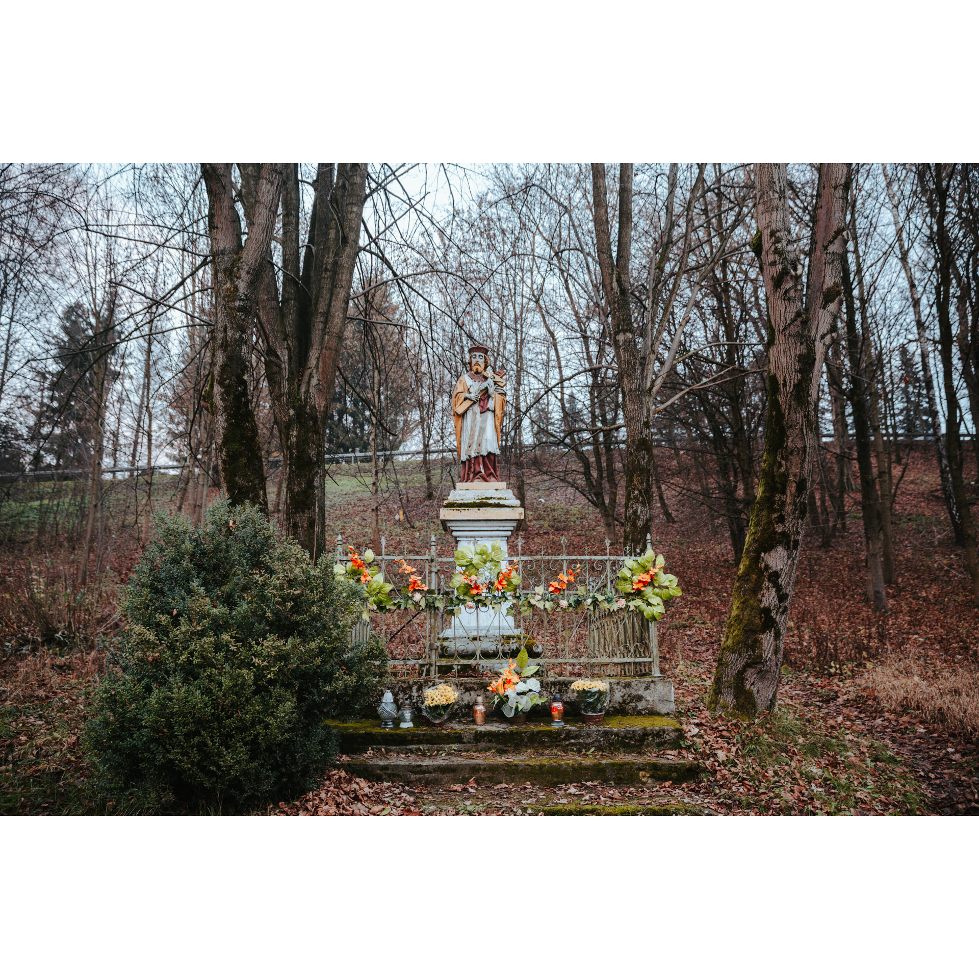Kapliczka z figurą świętego w lesie otoczona metalową barierką i kolorowymi kwiatami 