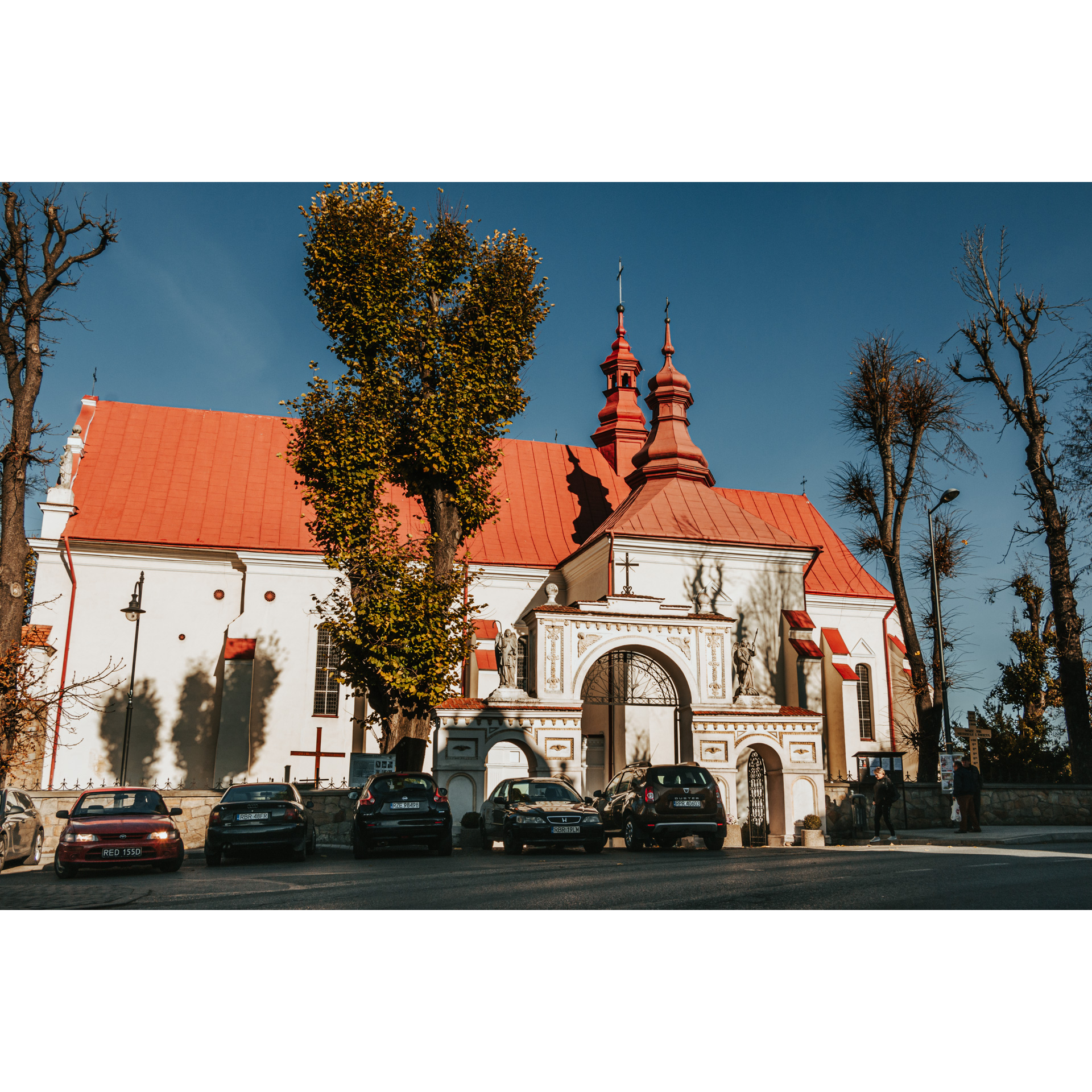 A bright brick church with a red roof and turrets and an entrance gate with ornaments and statues of saints