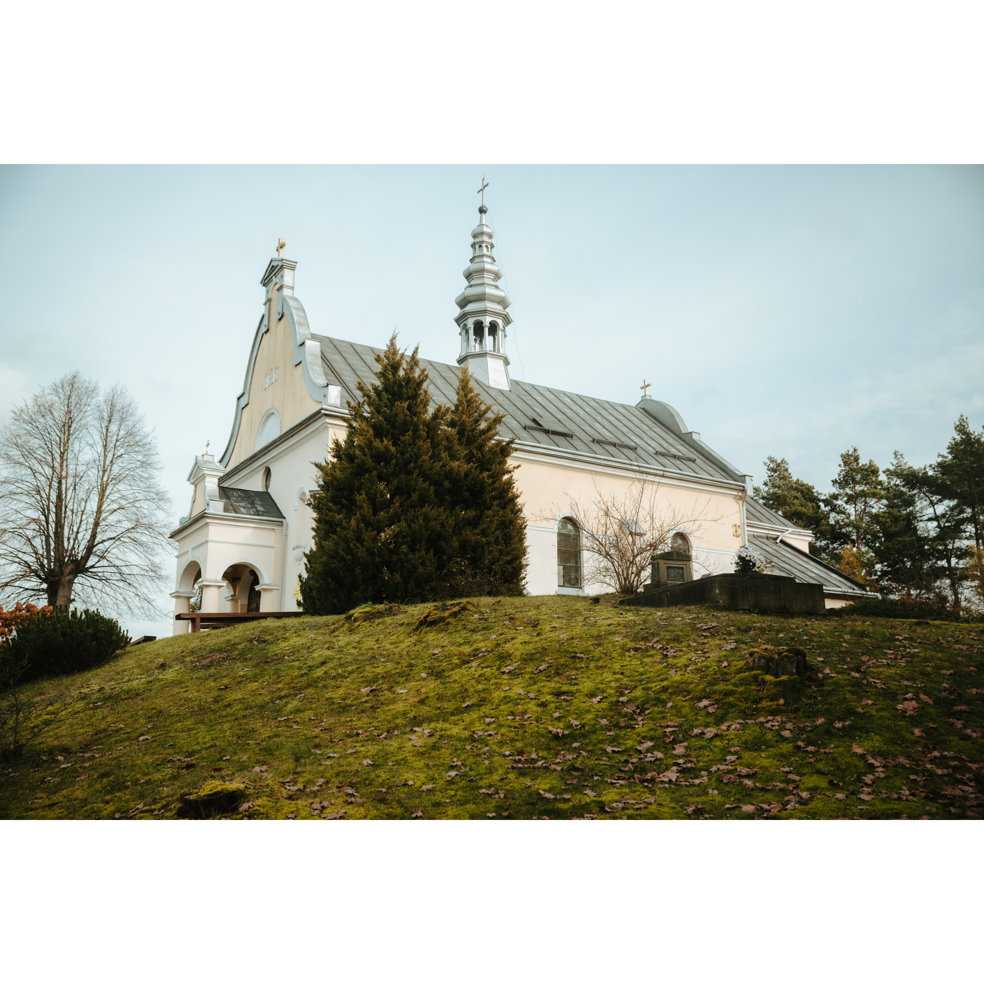 A bright brick church with a gray roof and a steeple on a grassy hill next to a dark green bush