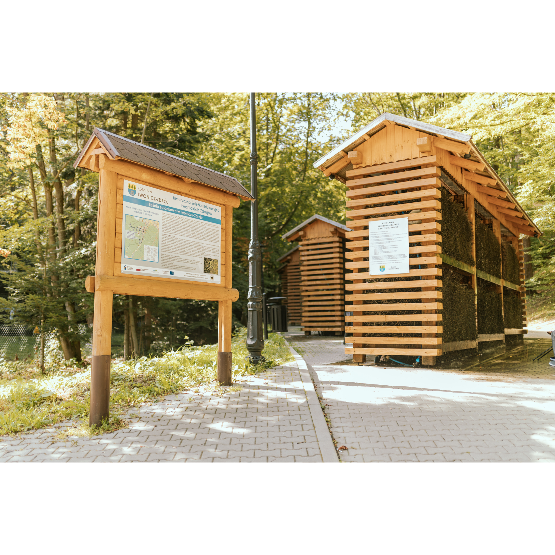 A wooden information board in the foreground describing the graduation towers behind it, i.e. four wooden structures 8 m long, 4 m high and 1 m wide, with a forest in the background