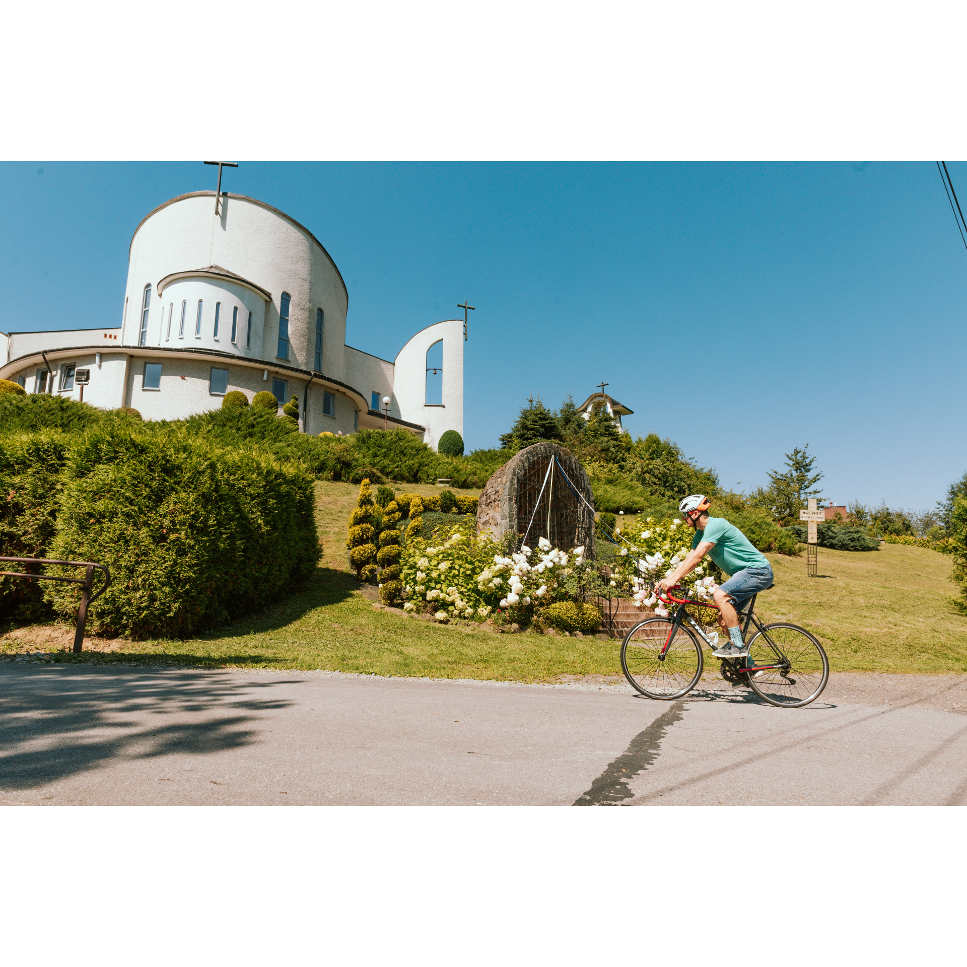 Na pierwszym planie rowerzysta w kasku jadący po asfaltowej ulicy, w tle na wzniesieniu biały budynek kościoła z okrągłą fasadą, wokół zielona roślinność i kapliczka