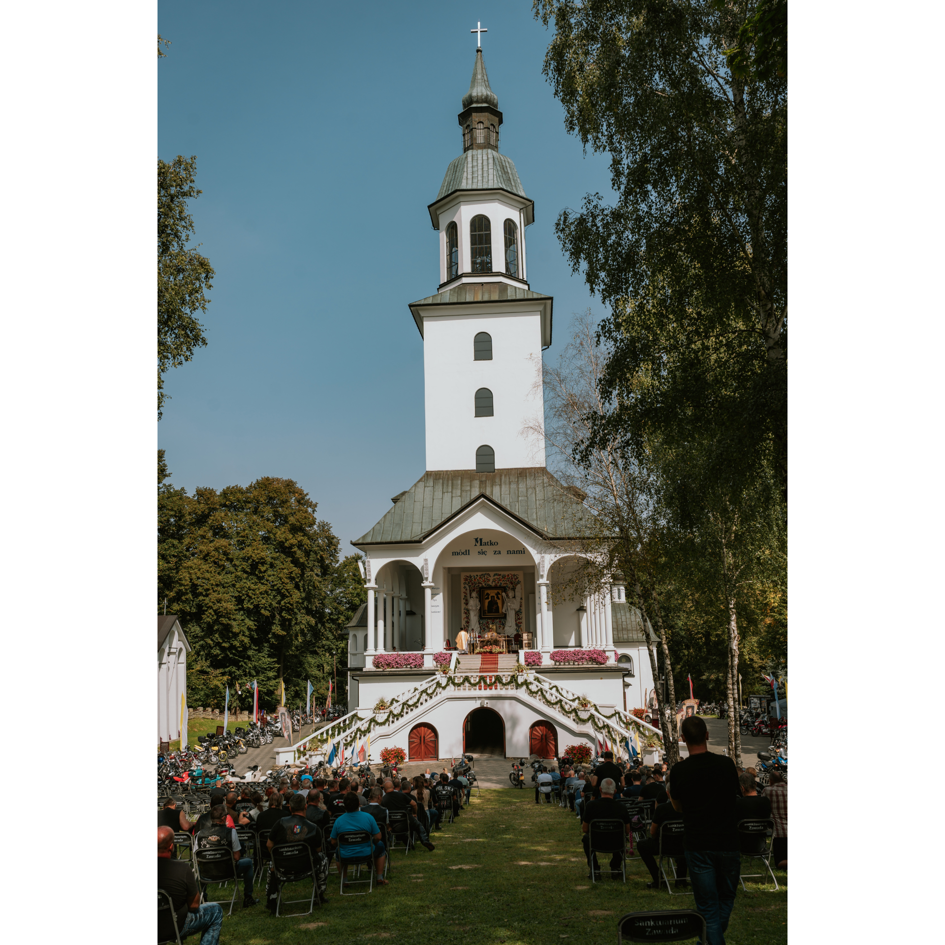 Widok na front strzelistego kościoła, na przodzie schody z obu stron prowadzące do wnętrza, na pierwszym tle ludzie siedzący na krzesłach, uczestniczący w nabożeństwie