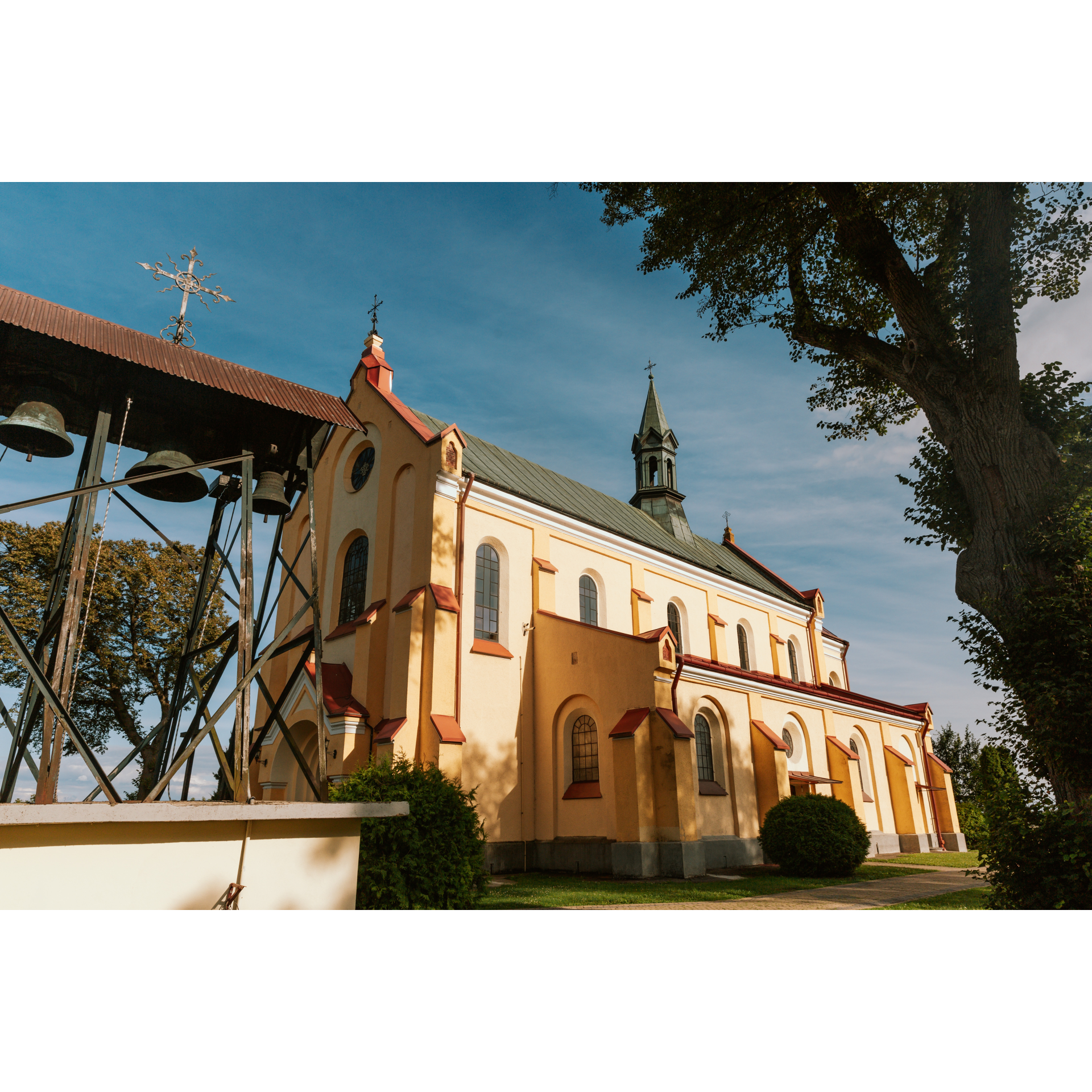 In the foreground a metal bell tower with three bells, in the background a tall, rectangular yellow church with a soaring turret