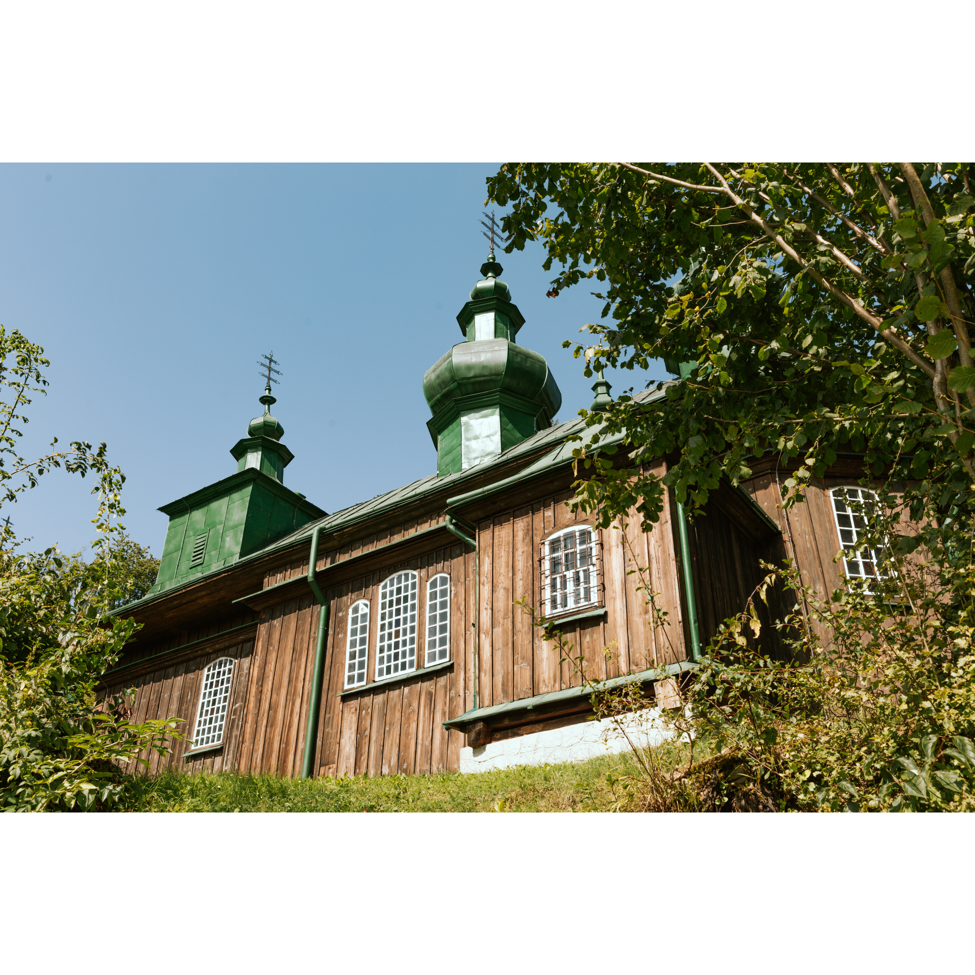 A wooden church with domed turrets and a green roof, green trees around