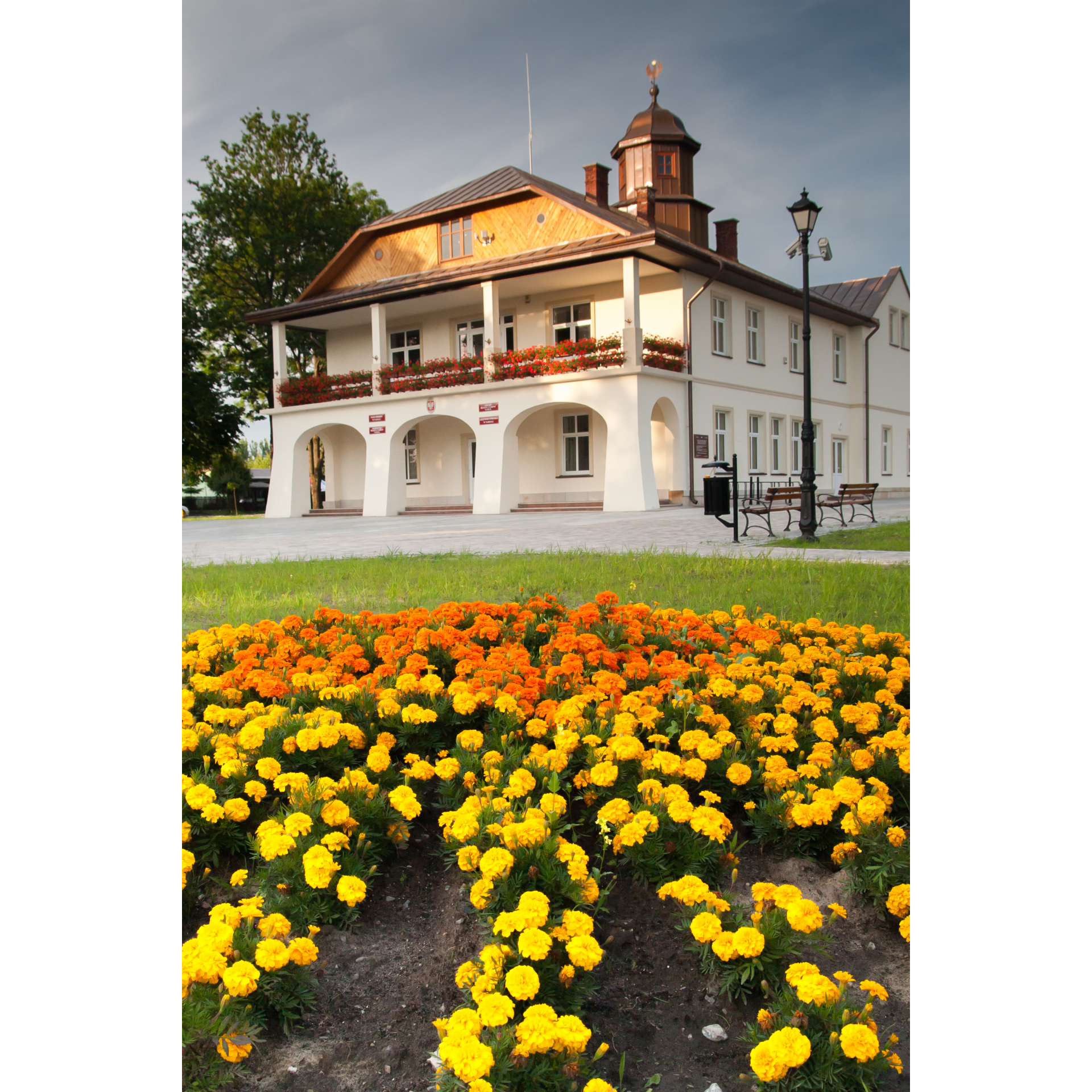 A bright building with arcades and an attic with a wooden finish, surrounded by yellow and orange flowers