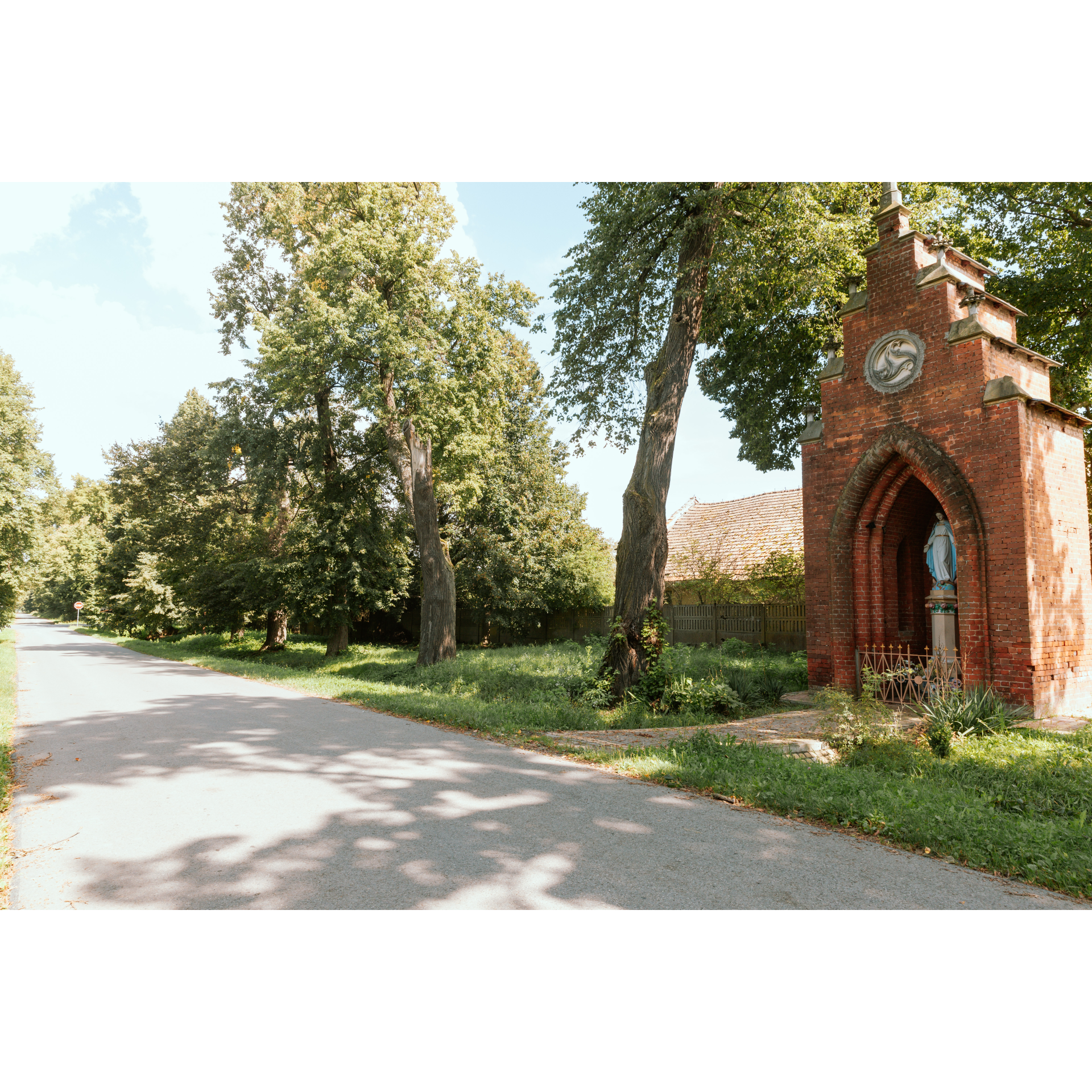 Stojąca przy drodze kapliczka zbudowana z czerwonej cegły o smukłej bryle z figurą Matki Boskiej 