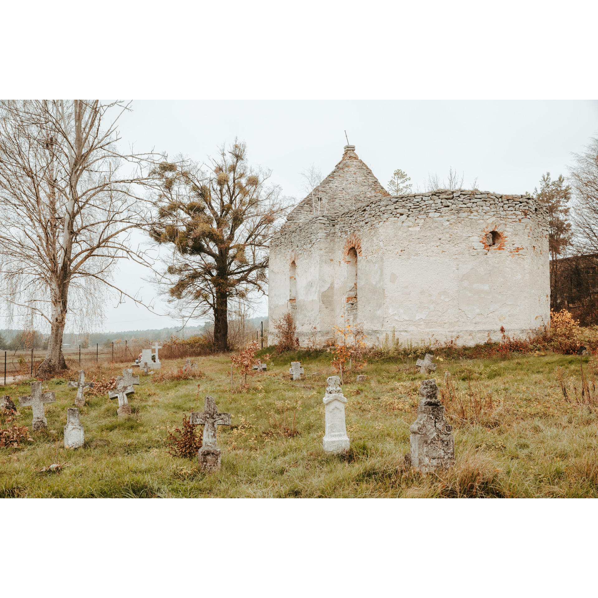 Biała, murowana ruina cerkwi z widocznymi kamiennymi krzyżami wokół na zielonej trawie