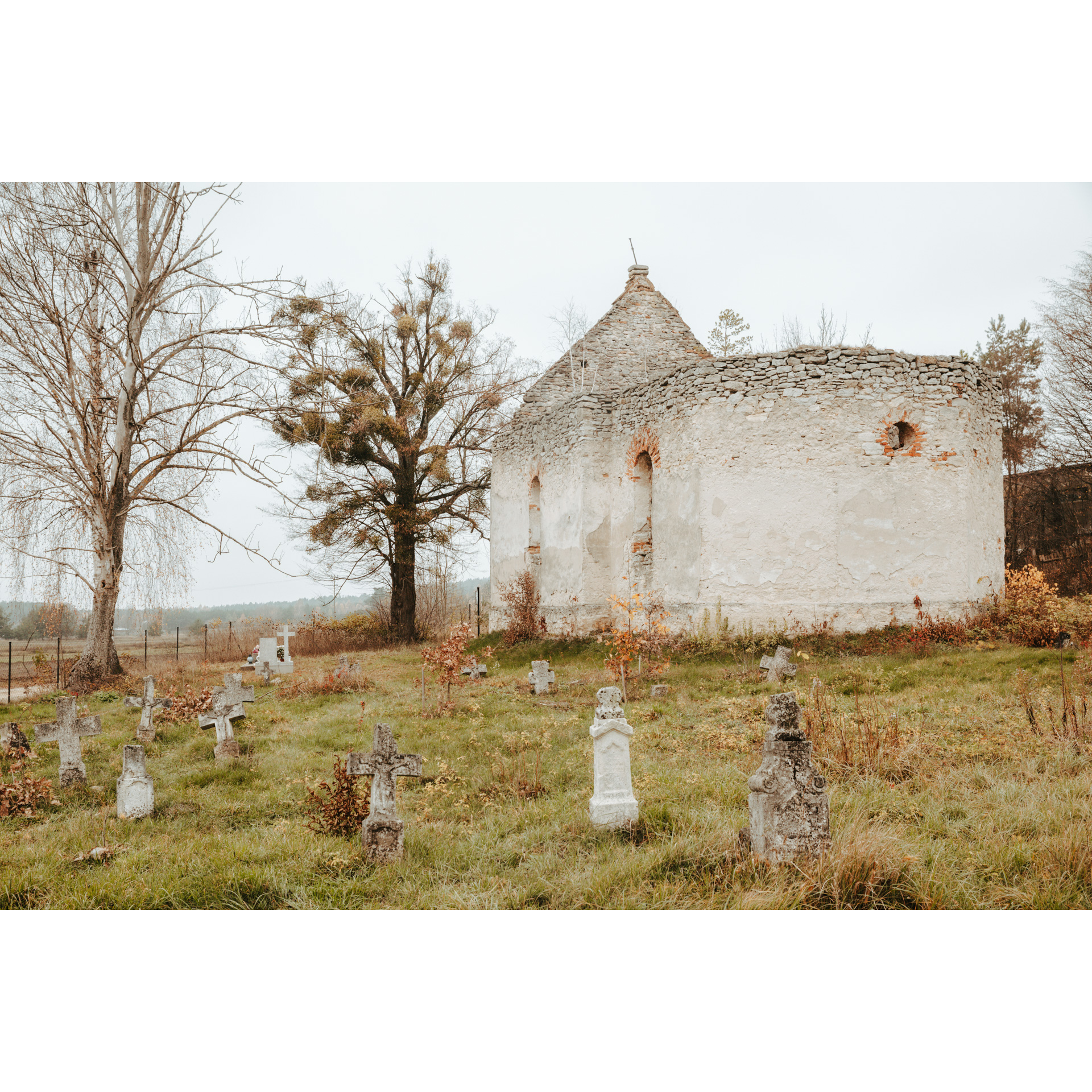 A white, brick ruin of an Orthodox church with visible stone crosses around it on the green grass