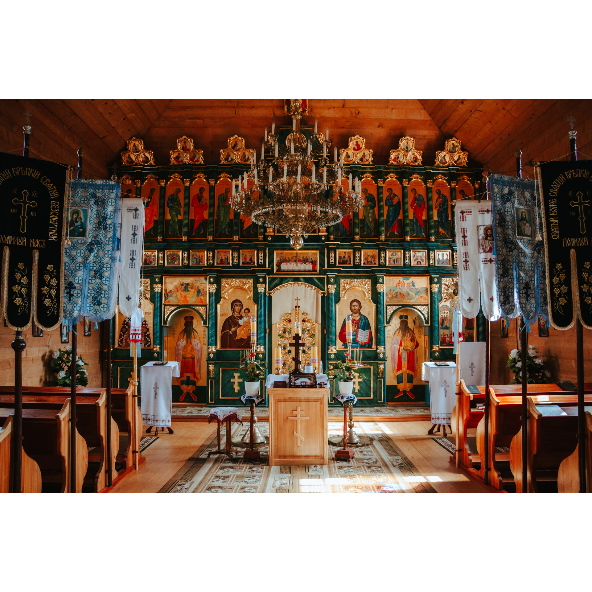 Ołtarz drewnianej cerkwi z ikonostasem, wiszącym, kryształowym żyrandolem i drewnianymi ławkami po bokach 