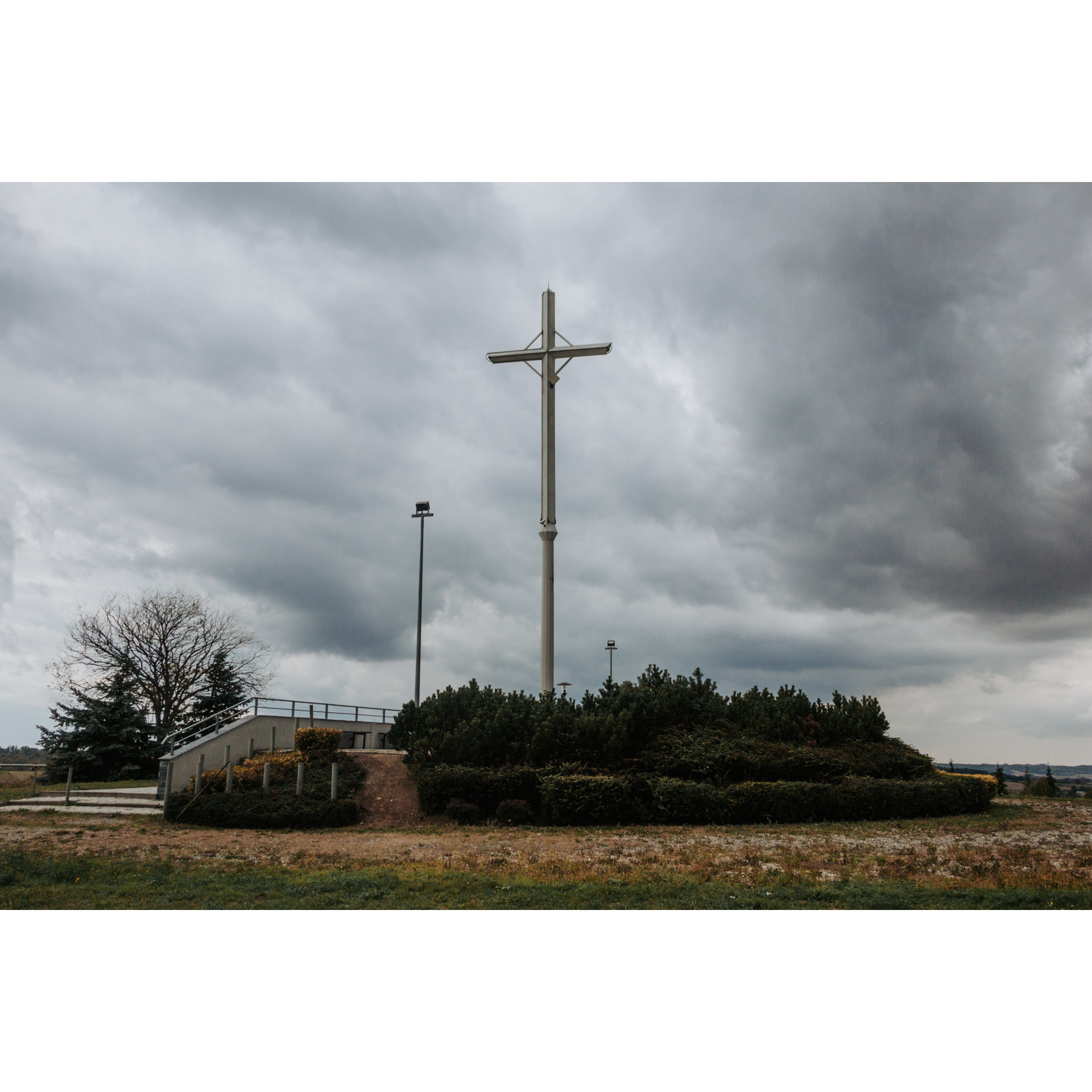 A tall cross surrounded by low bushes against a cloudy gray sky