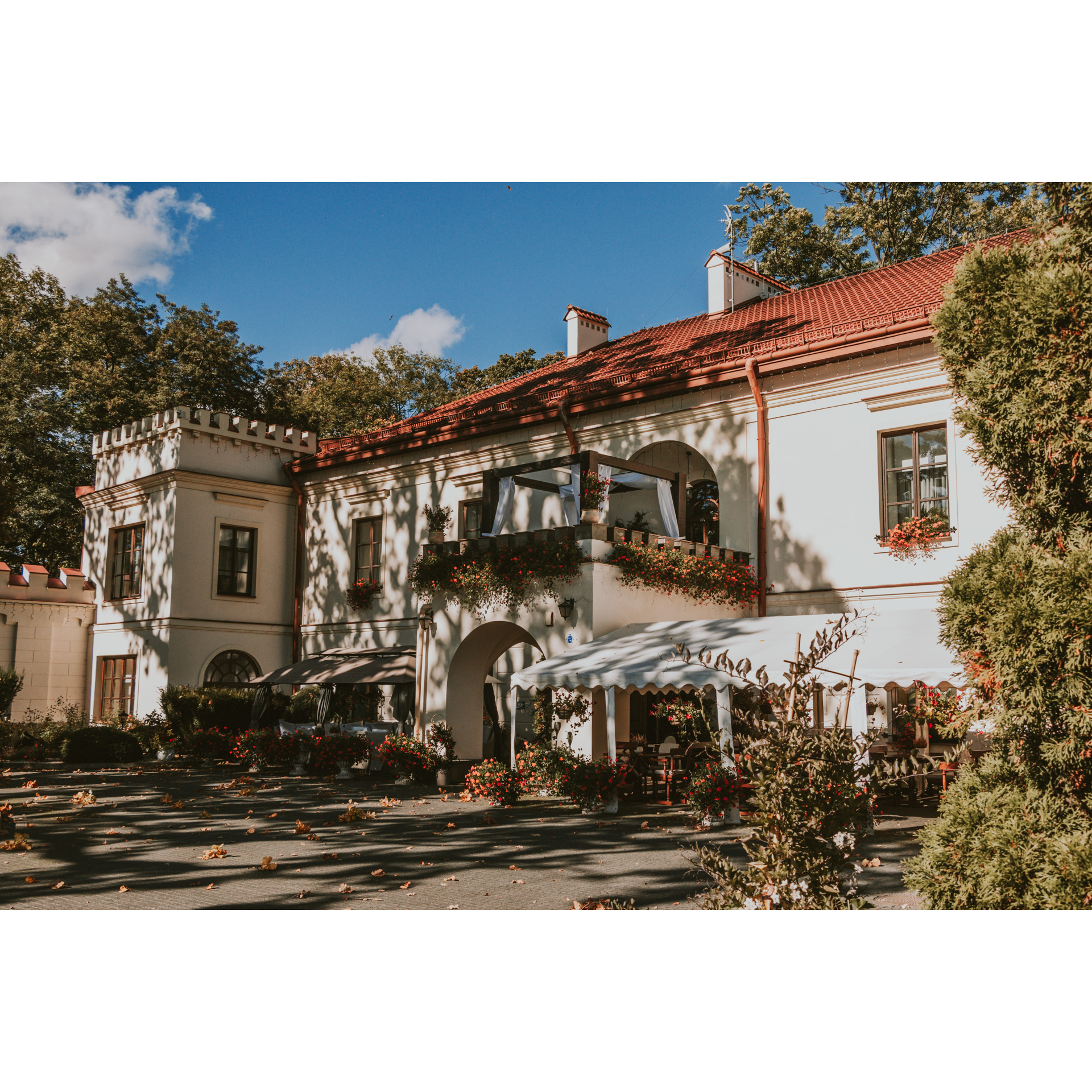 A bright manor house with a red roof and a balcony among trees with numerous rose compositions