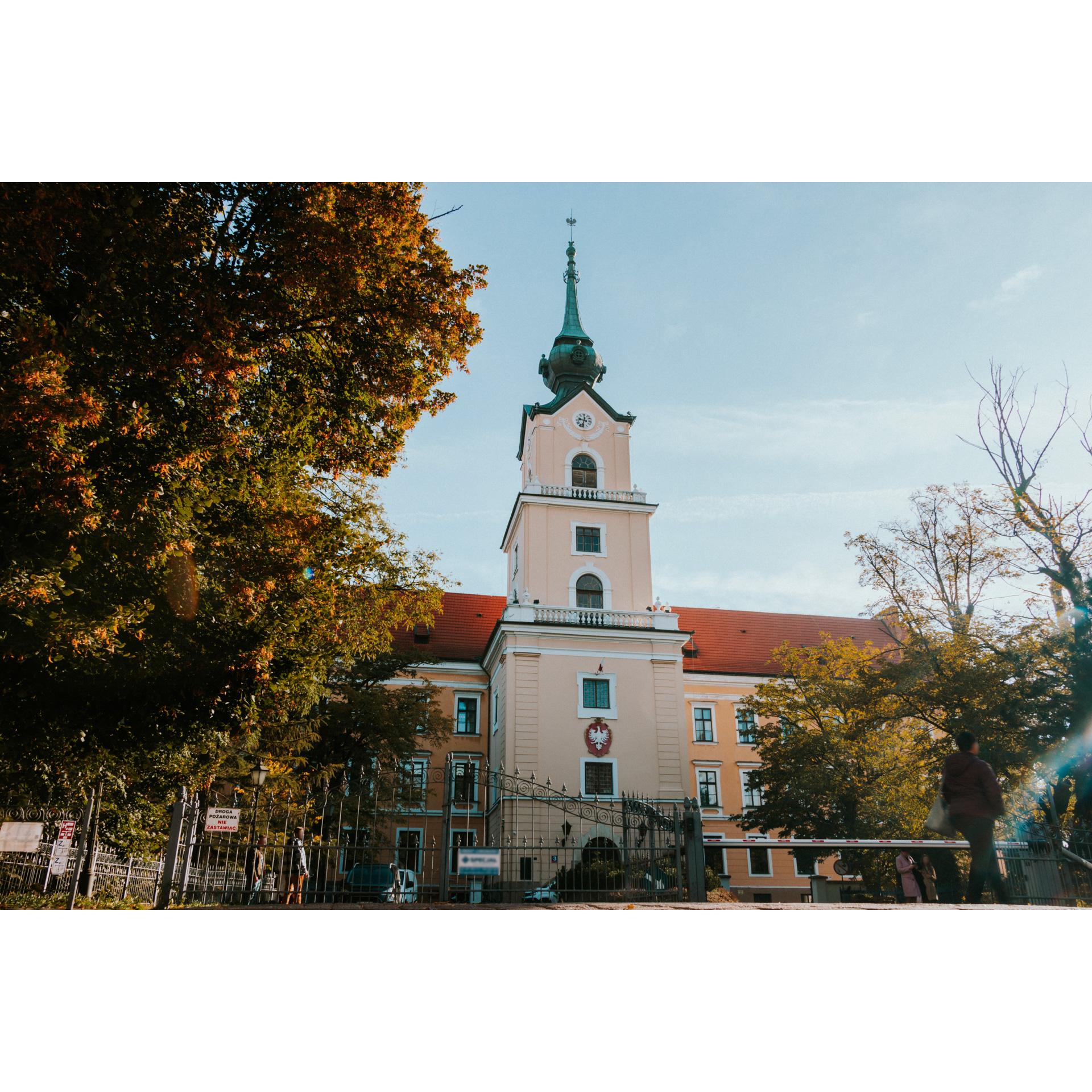 Wieża zamku z zegarem i godłem Polski zakończona zieloną wieżyczką wśród drzew