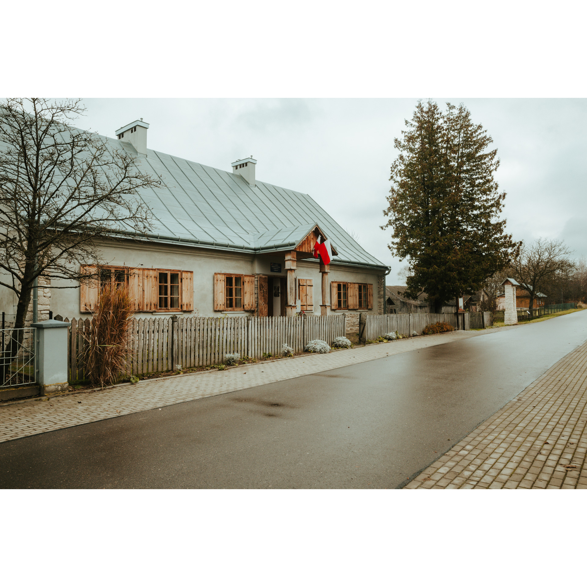 Murowany niski dom z drewnianymi okiennicami, flagą Polski i szarym dachem otoczony białym, drewnianym płotem przy asfaltowej drodze z chodnikiem
