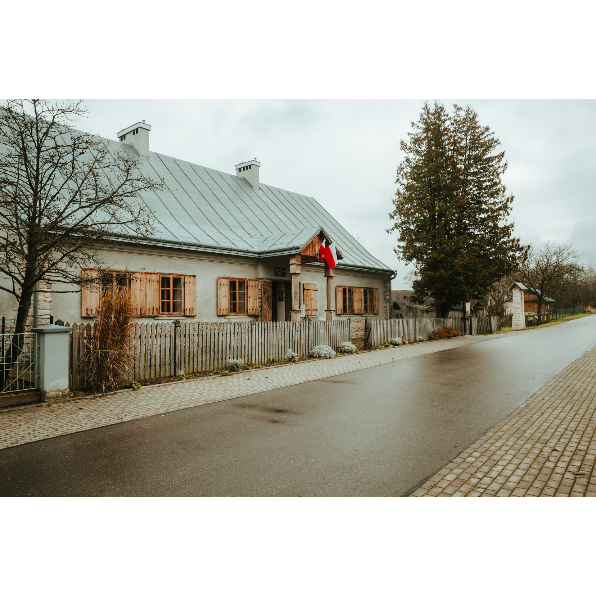 A low-rise brick house with wooden shutters, a Polish flag and a gray roof surrounded by a white wooden fence by an asphalt road with a sidewalk