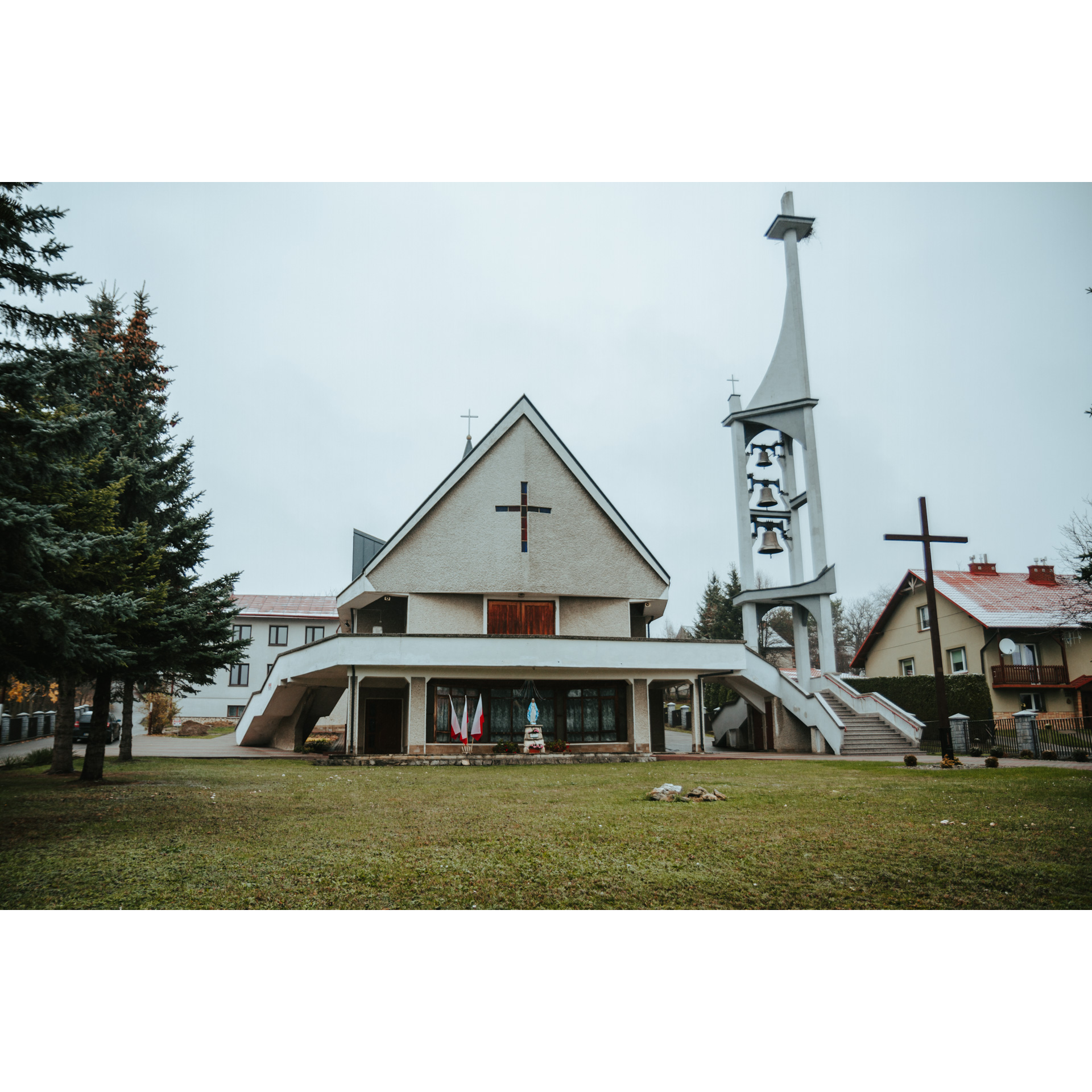 Kościół z trójkątną fasadą i krzyżem i figurą Matki Bożej pod schodami obok wysokiej dzwonnicy z trzema dzwonami