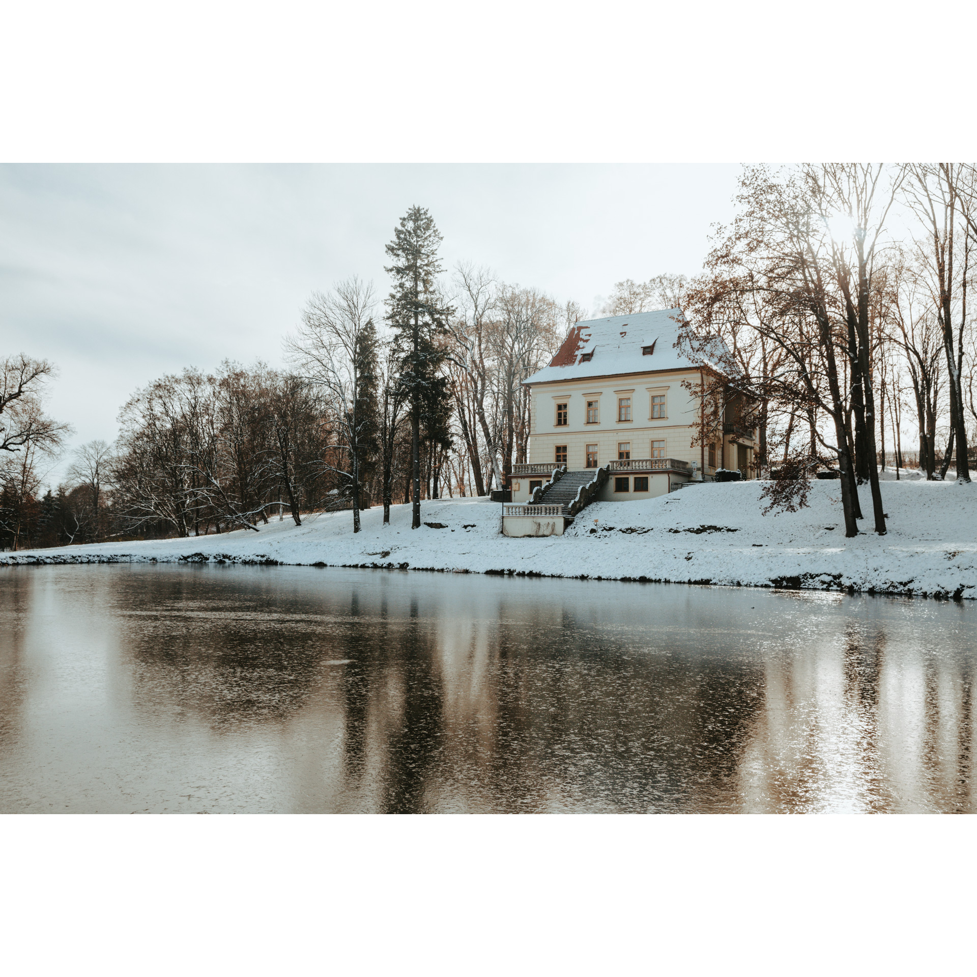 Light beige country house with a snowy roof with wide stairs leading to the snowy shore of a pond