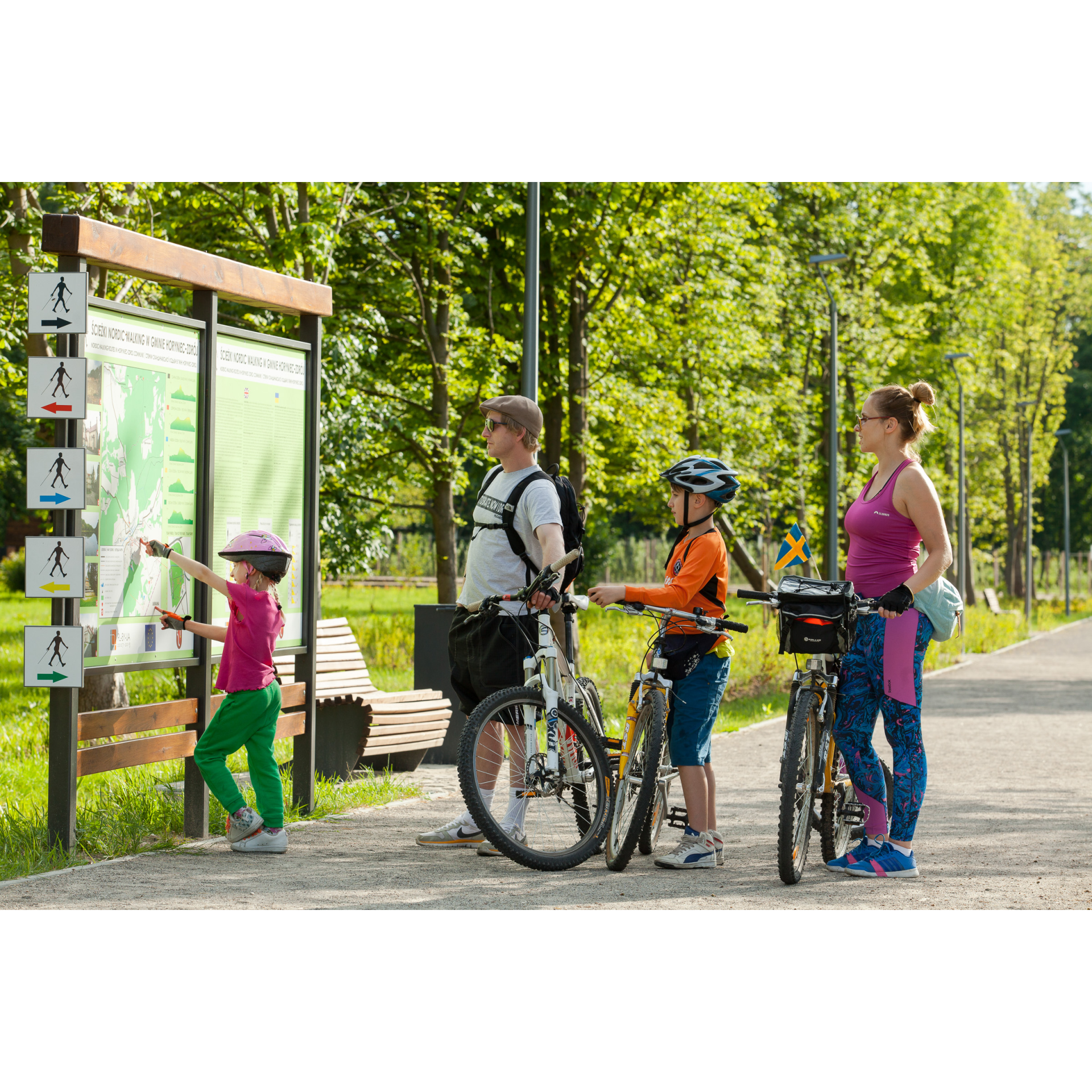 Rodzina z dziećmi przy tablicy informacyjnej w parku, przygotowująca się do wycieczki rowerowej.