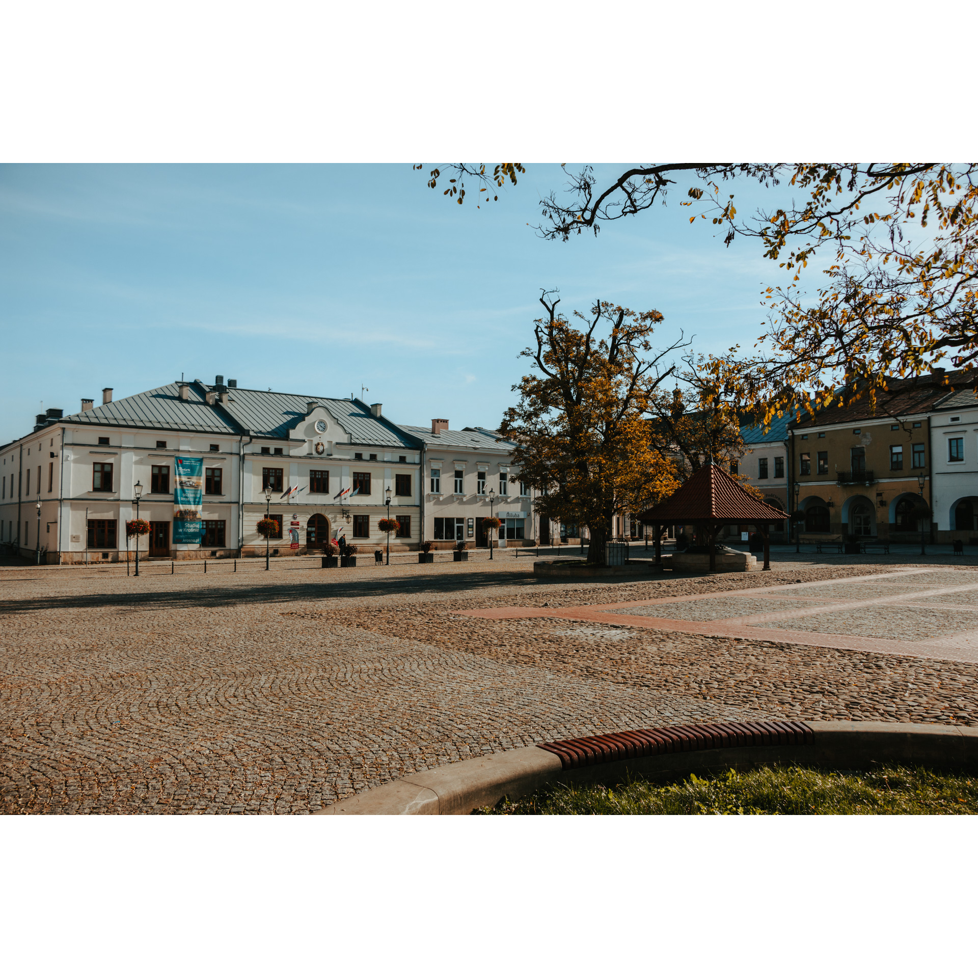 Plac rynkowy z widokiem na białą, dużą kamienicę, zabudowania miejskie oraz studnię stojącą przy jesiennym drzewie