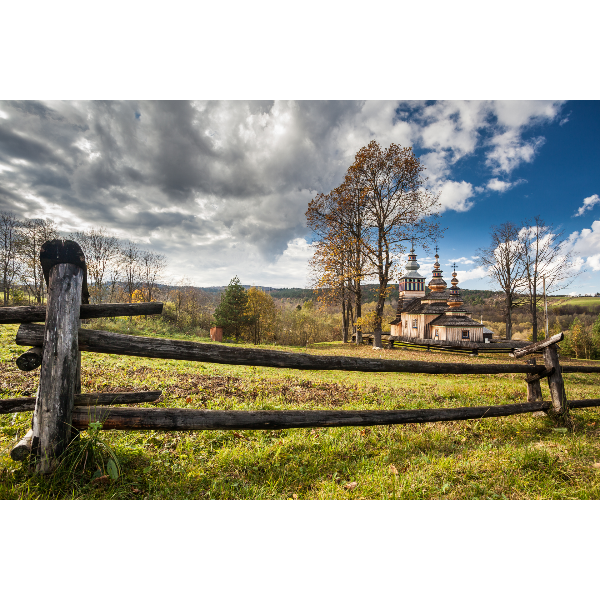 A view of a wooden Orthodox church surrounded by a wooden fence against the backdrop of forested hills