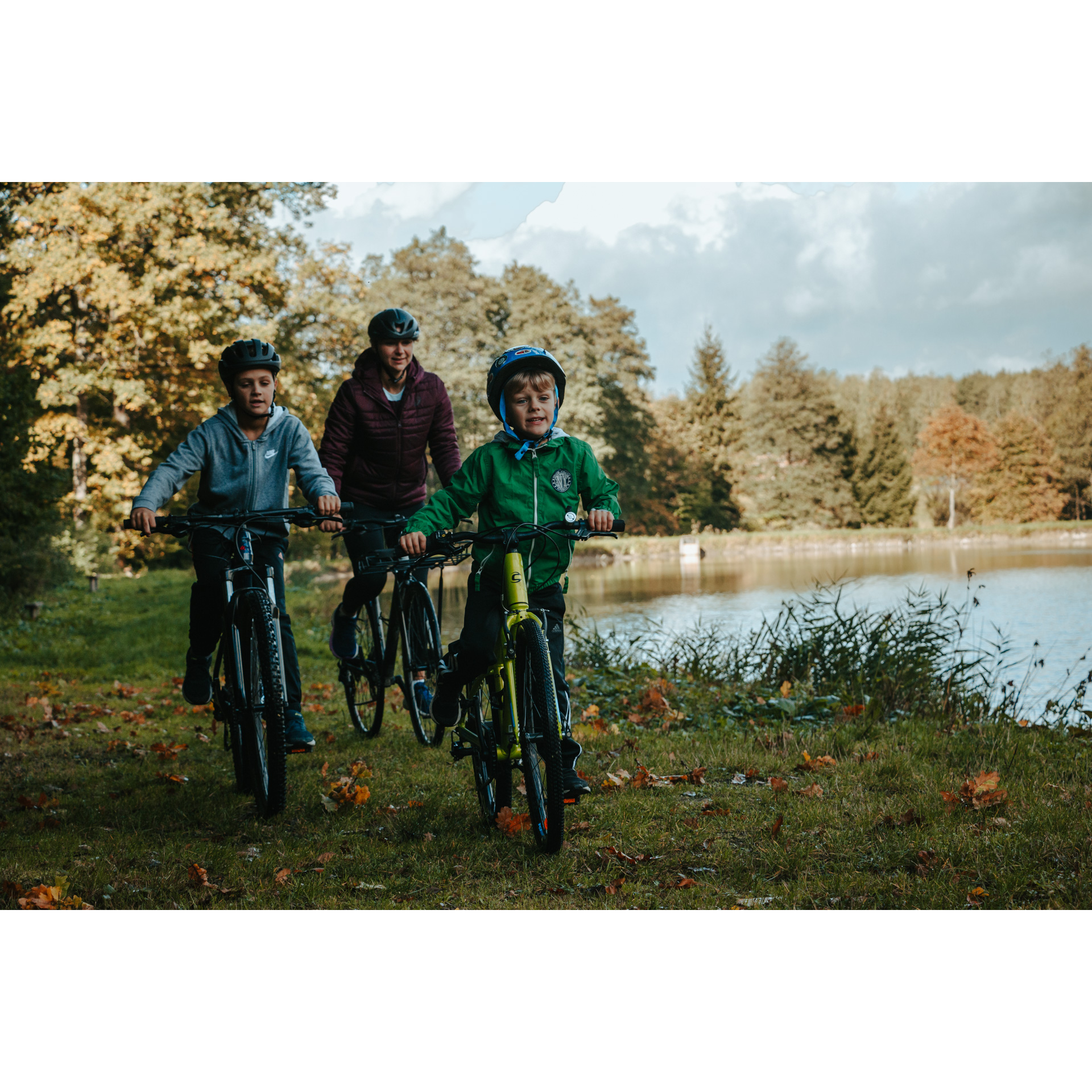 Two boys and a woman cycling on the grass along the water, with trees in the background