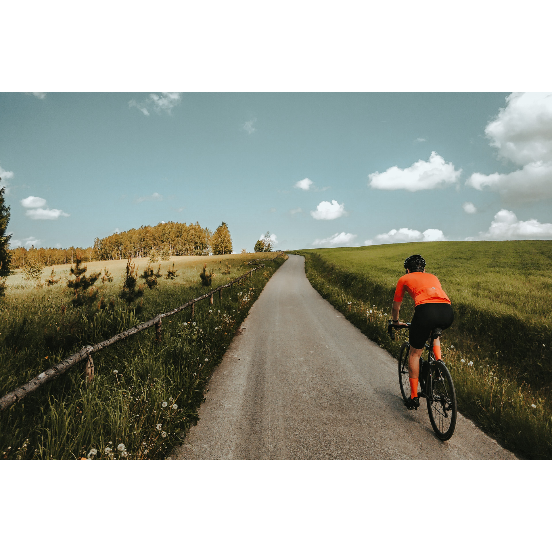 Rowerzysta w pomarańczowej jadący po szutrowej drodze pomiędzy wysoką trawą odgrodzoną po lewej stronie płotem 