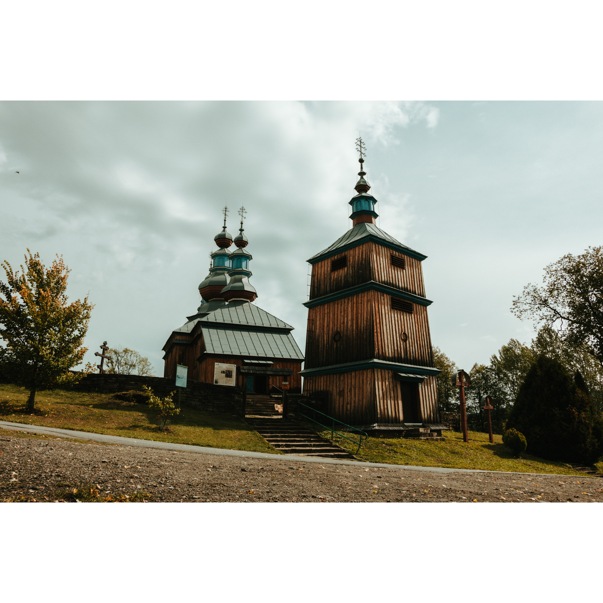 A wooden church with a large, high bell tower and domes, accessed by stone stairs with a green railing