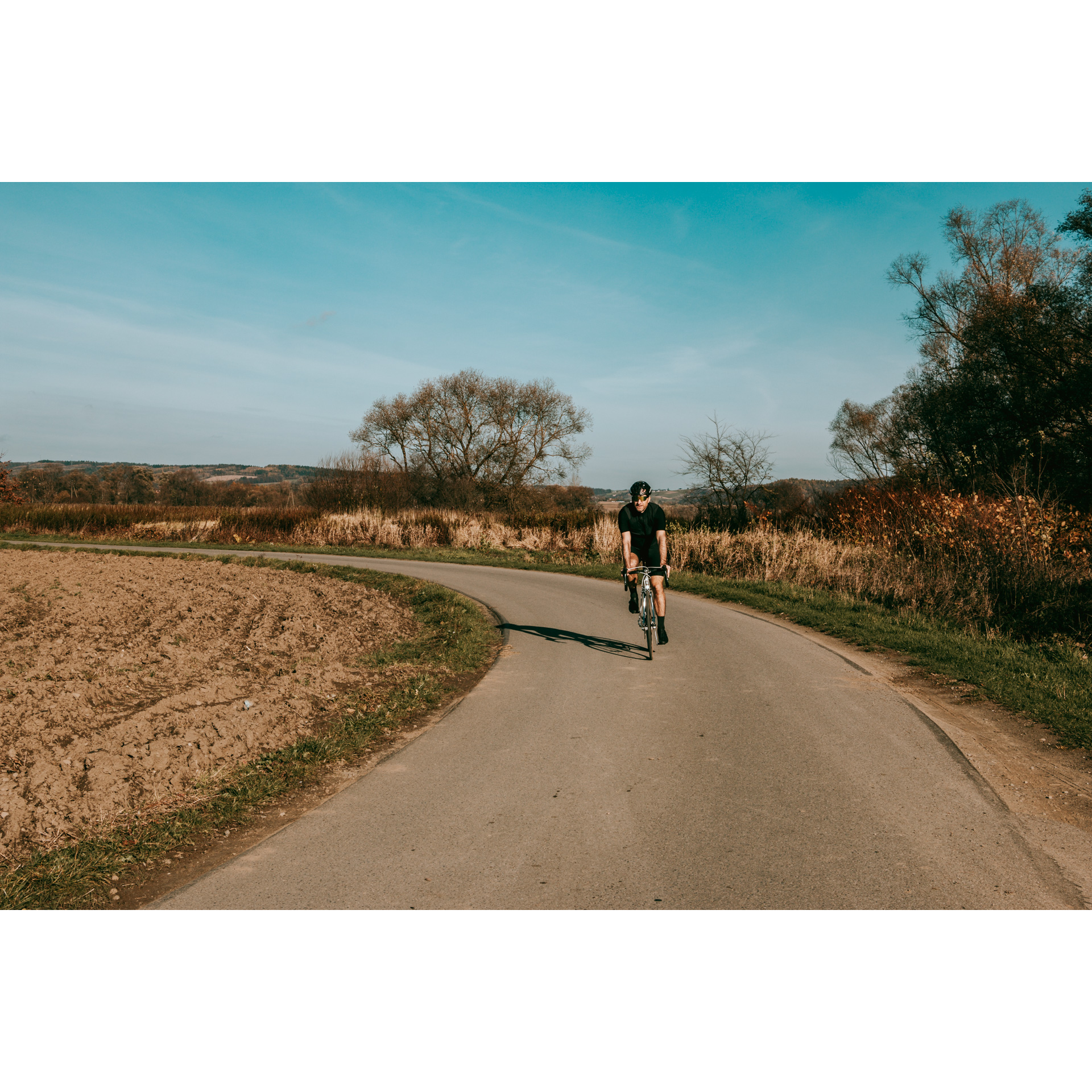 Rowerzysta w czarnym stroju jadący drogą asfaltową mijający pole orne i beżową roślinność