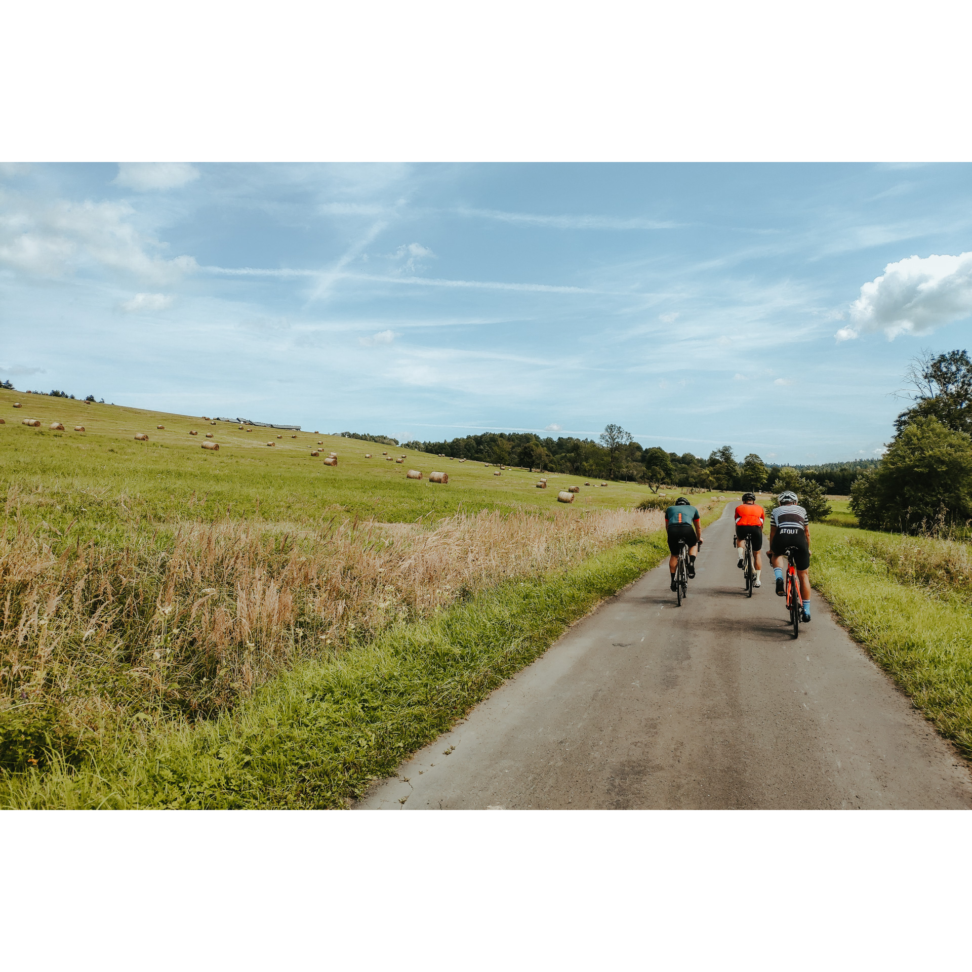 Three cyclists in colorful clothes riding an asphalt road surrounded by meadows with bales of hay