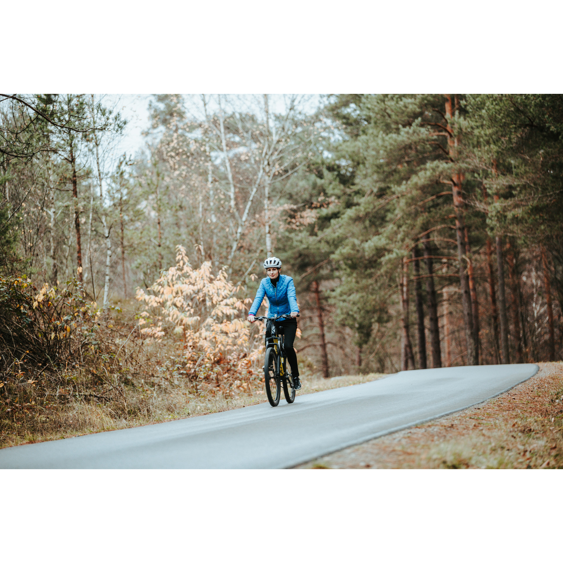 Rowerzystka w niebieskiej kurtce i białym kasku jadąca drogą asfaltową prowadzącą przez las
