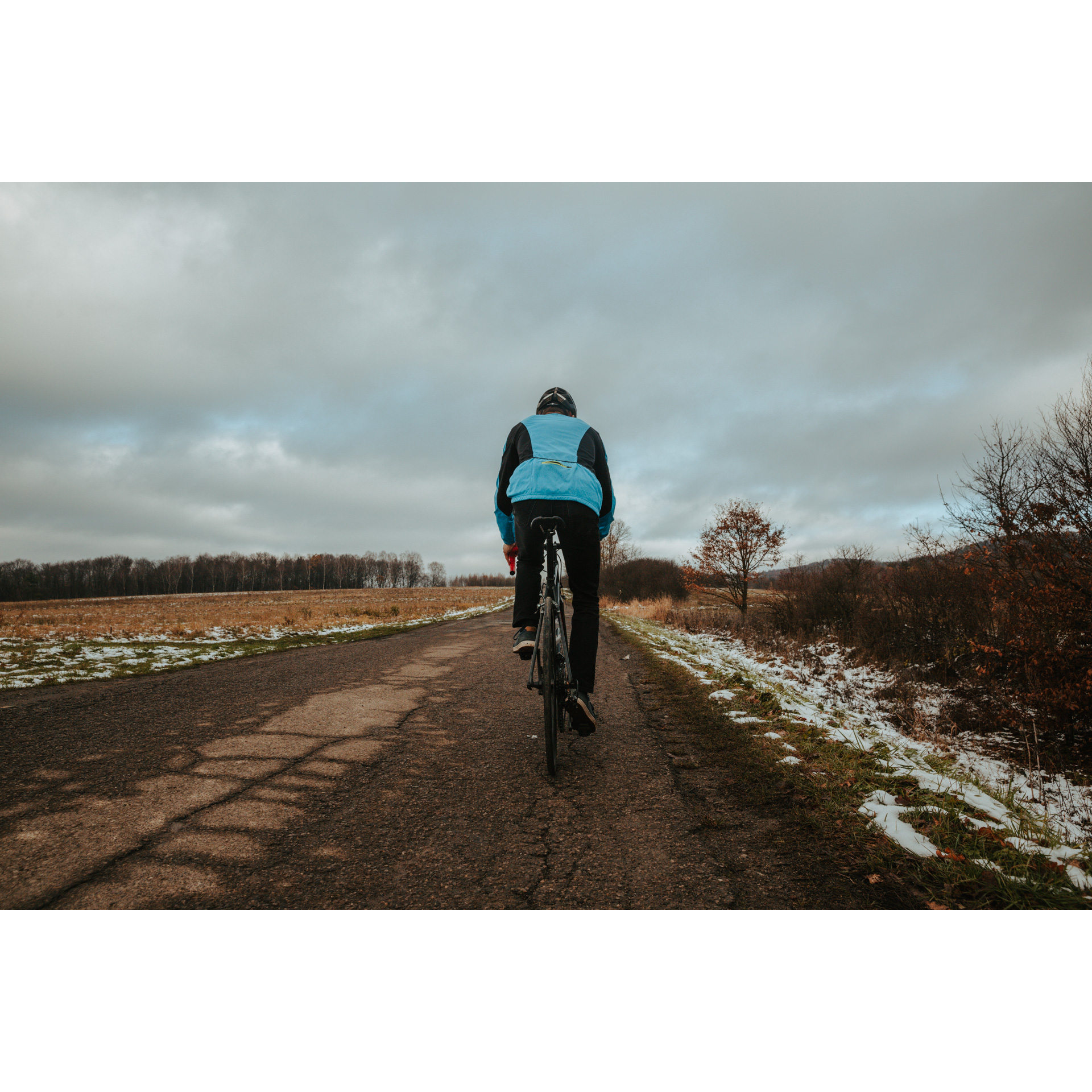 Rowerzysta w niebieskiej kurtce i czarnym kasku jadący drogą asfaltową w otoczeniu jesiennej polany