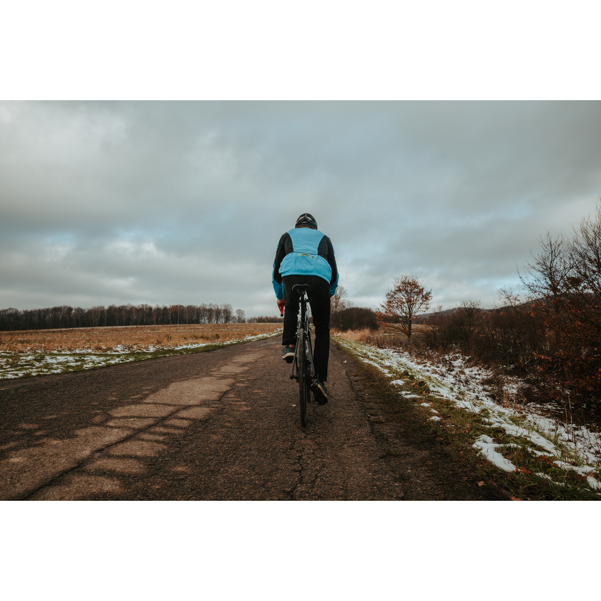 A cyclist in a blue jacket and a black helmet riding an asphalt road surrounded by an autumn glade