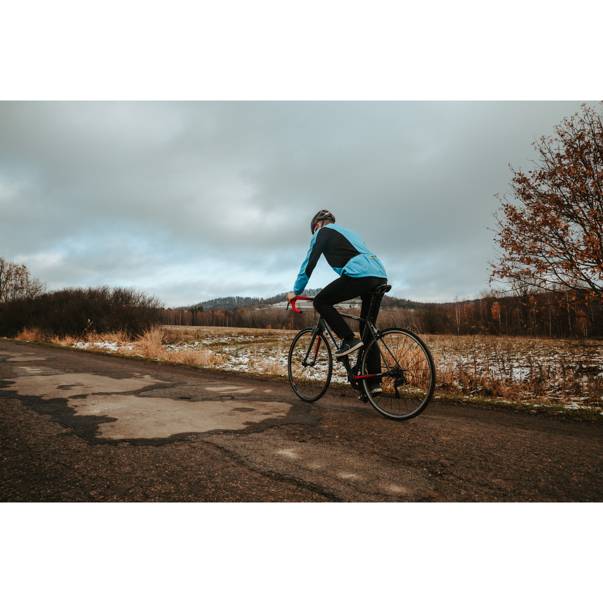 Rowerzysta w niebieskiej kurtce i czarnym kasku jadący drogą asfaltową w otoczeniu jesiennej polany