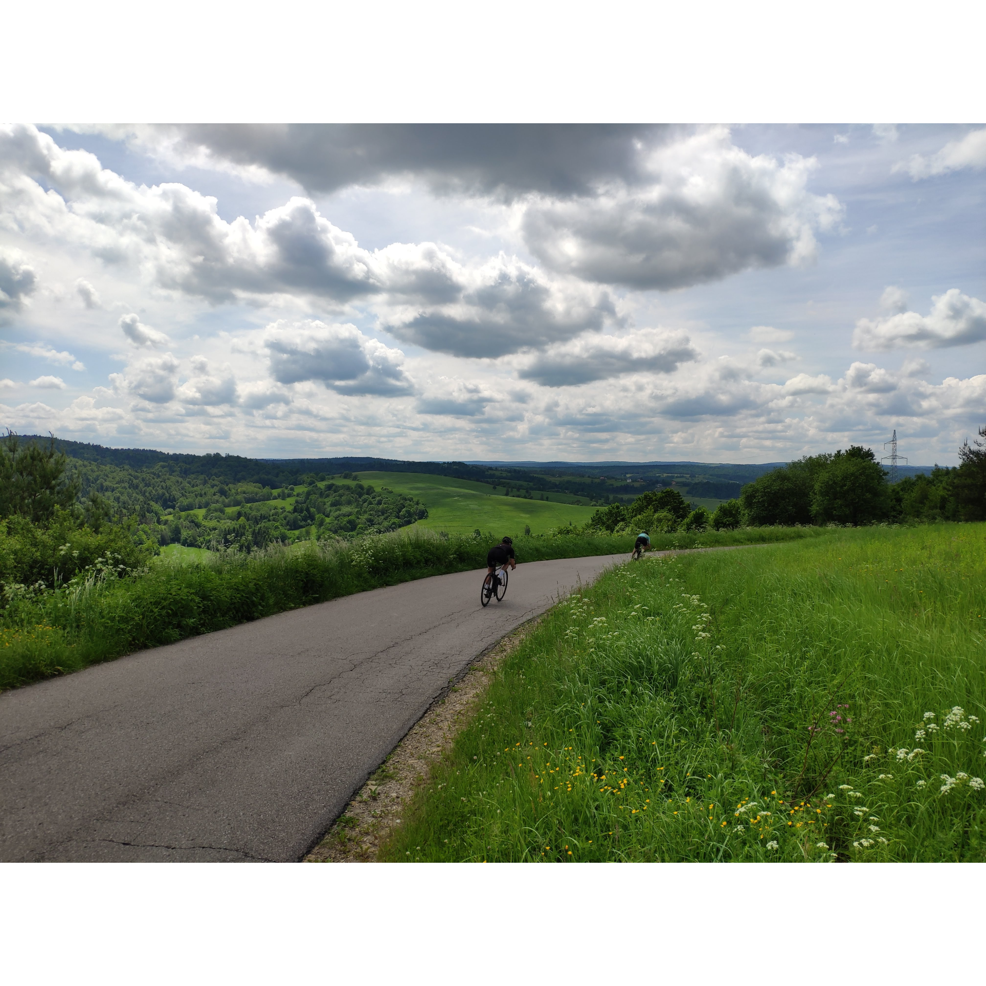 Dwójka rowerzystów w strojach kolarskich jadąca na rowerach drogą asfaltową prowadzącą przez łąki na tle wzgórz