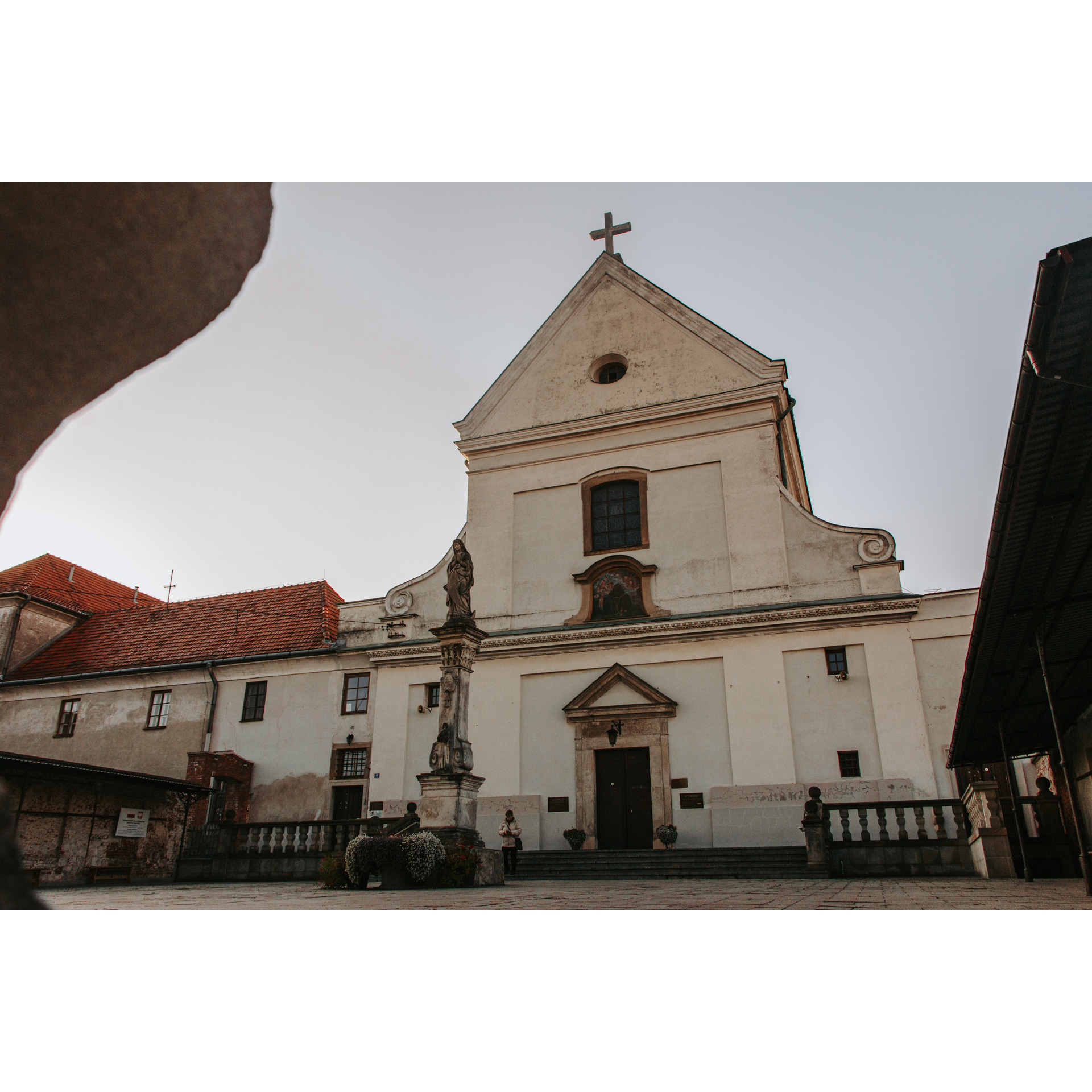 Jasny, wysoki kościół z trójkątnym frontem i krzyżem na szczycie oraz figura Matki Boskiej na postumencie przed budynkiem